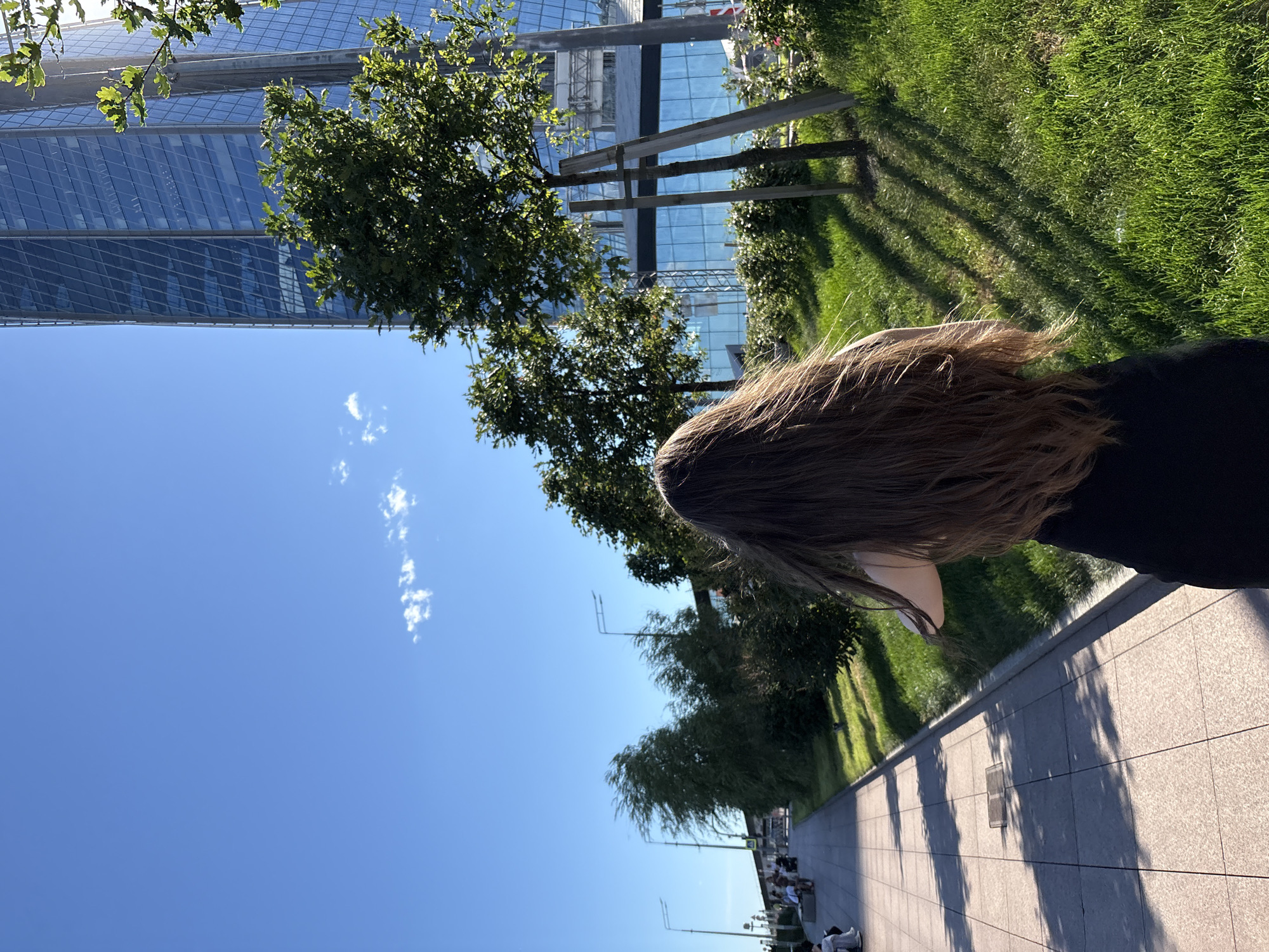The image captures a scene on a sunny day, likely late afternoon given the long shadows cast on the paved walkway. A person's long, light brown hair dominates the foreground, suggesting they are walking away from the camera. The background features a modern building with a glass facade, interspersed with green trees and a well-maintained lawn. The sky is a clear, vibrant blue with a few wispy clouds. In the distance, along the walkway, several people are seated on benches and walking, indicating a public space. The overall atmosphere is serene and pleasant, with the sunlight creating a warm glow. The scene is set in a location that appears to be urban, with the architectural style of the building and the presence of trees and park-like landscaping. Although the exact location is not explicitly stated within the image, the prompt provides context that it is Saint Petersburg, Russia.