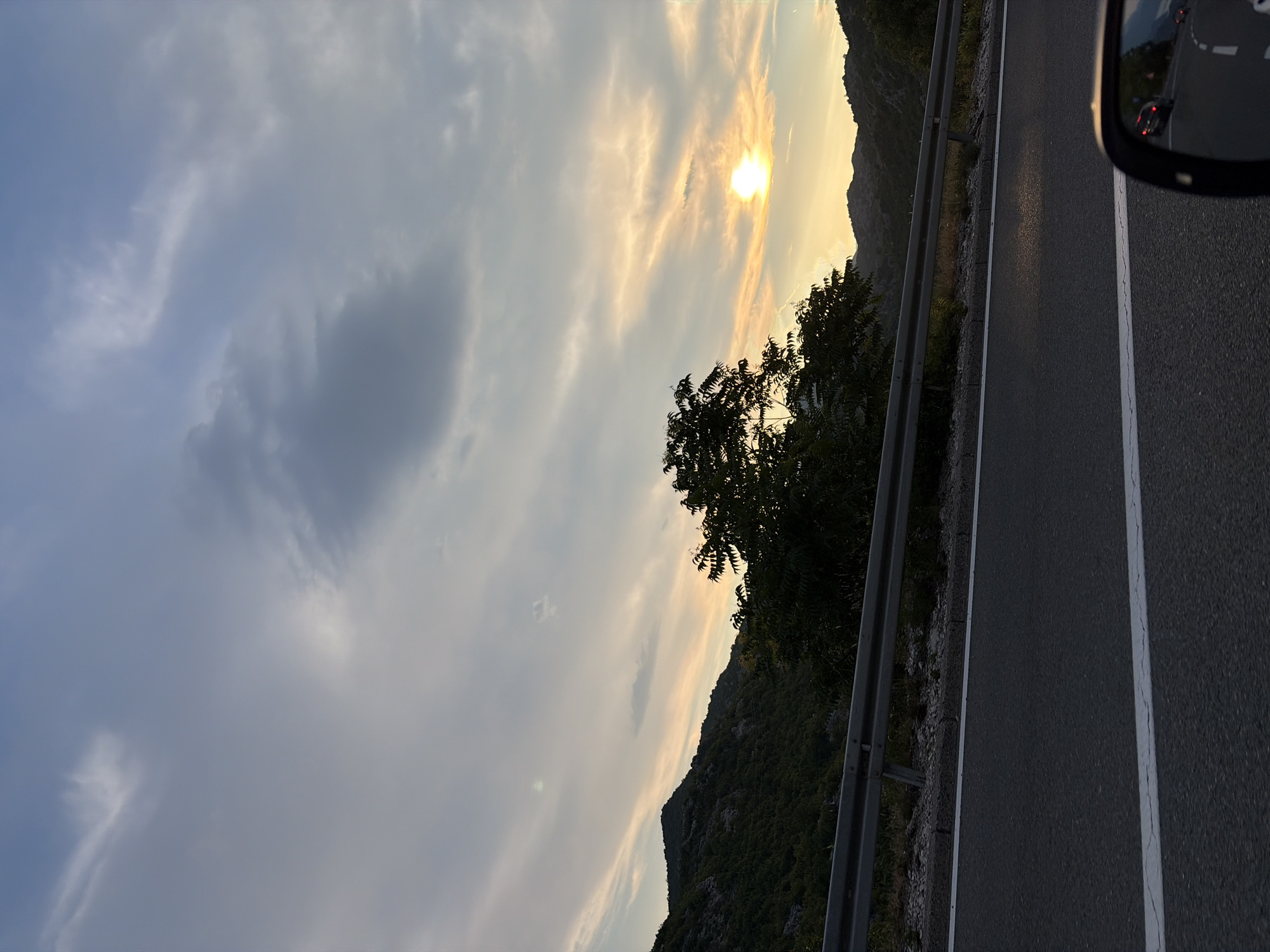 This media file depicts a view from a vehicle driving along a road in Gornja Brova, Montenegro. The scene captures a mountainous landscape bathed in the warm light of a setting sun. The sky is a mix of soft blue and wispy, golden clouds, with the sun partially obscured by them, creating a beautiful twilight effect.
On the left side of the frame, a lush, green mountainside slopes upwards, dotted with trees and vegetation. A metal guardrail runs along the edge of the road, separating it from the steep incline. On the right, the asphalt road is visible with a white line marking the lane. A side mirror of the vehicle is visible in the upper right corner, reflecting the road ahead and other vehicles.
The time of day is clearly evening, indicated by the sunset and the softer light. The weather appears to be clear with scattered clouds. The overall mood is serene and tranquil, with a sense of journey and natural beauty. There is no visible text in the image.