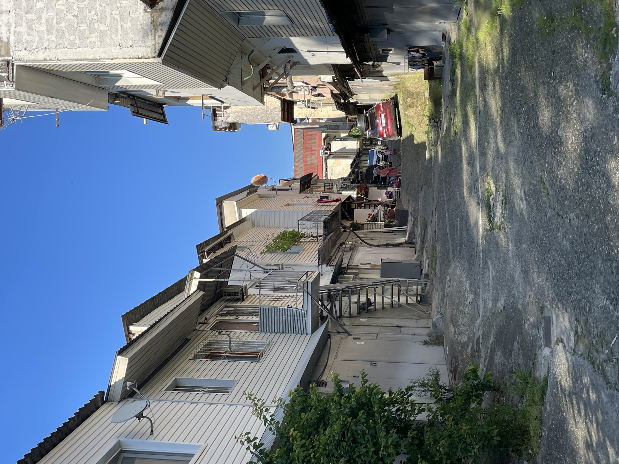The image presents a steep, narrow residential alley in Sochi, Russia, viewed under a clear, cloudless blue sky. The alley ascends from the foreground, flanked by multi-story buildings on both sides.
On the left, structures feature light-colored horizontal siding and corrugated metal roofs. Multiple windows are visible, some equipped with security bars. A white satellite dish is affixed to the exterior of one building. An external metal staircase, incorporating a small landing, provides access to an upper floor, while the ground level appears to consist of garage or storage units with closed doors. A large, dense green bush occupies the lower-left foreground.
Buildings on the right side display varied construction, including sections of exposed concrete block and areas with light-colored siding or plaster. Multiple awnings and extensions protrude from the facades. A clothes drying rack with garments is positioned near the lower-right foreground.
The alley's surface is uneven, consisting of cracked and patched asphalt or concrete, with scattered patches of grass and weeds growing through the fissures. Sunlight casts distinct shadows across the ground and building surfaces. In the mid-ground, an adult figure in a red garment and a child in light clothing are visible near the left side of the alley. Further up the incline, a red SUV, fitted with a roof box, is parked facing uphill on the right side of the alley, partially obscuring a white vehicle positioned behind it. Air conditioning units are mounted on the exterior walls of several buildings.