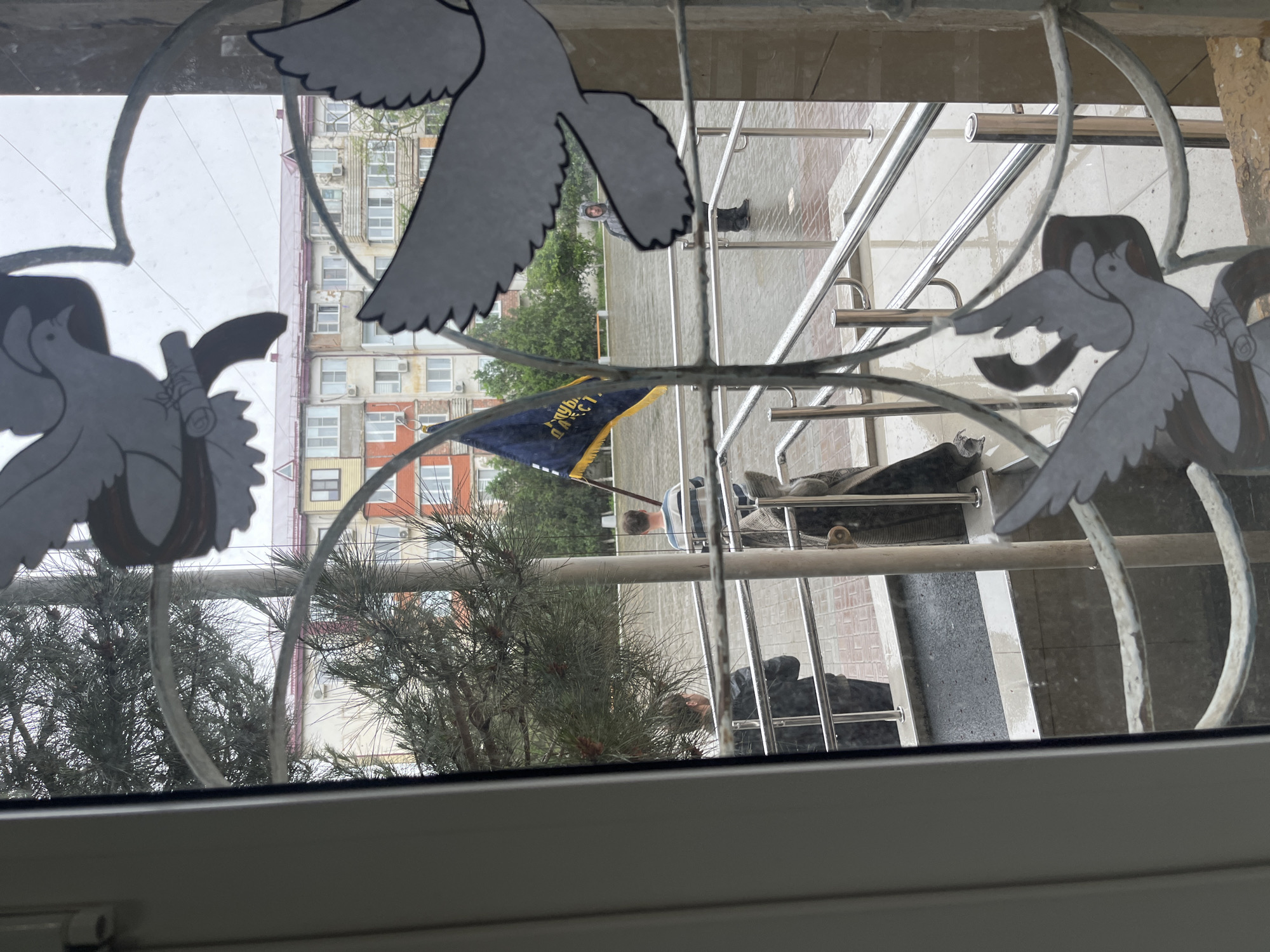 A view through a window pane decorated with metallic dove outlines. The outside scene, apparently in Kaspiysk, Russia, includes a multi-story apartment building with rows of windows and a blue flag. A person can be seen near a railing. There are also trees and greenery visible. The scene is viewed from indoors.