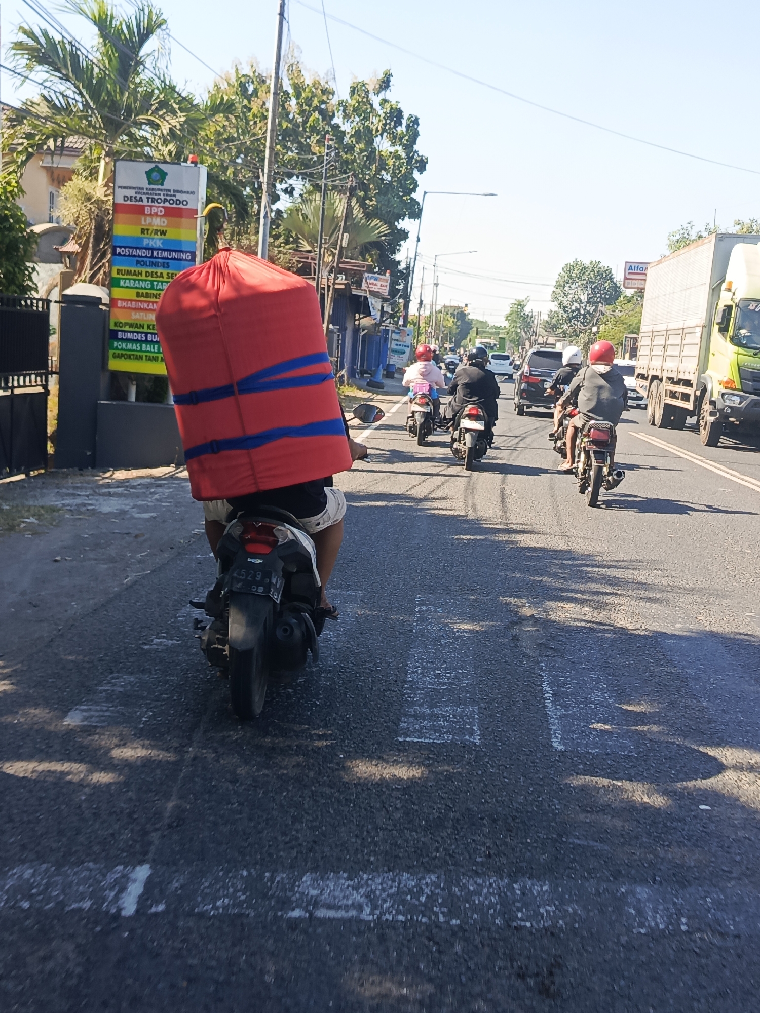 In Krian, Indonesia, a man rides a motorcycle down a street, carrying a large, red, cylindrical object wrapped with blue straps. The object is significantly taller than the rider. Several other motorcycles are visible on the road, with riders wearing helmets. A light-colored truck with a trailer is in the right lane, and a dark-colored SUV is in the left lane. A sign is visible on the left side of the road, with text in Indonesian, indicating a village name "DESA TROPODO" and other civic organizations. The sky is clear and blue, and the sun is casting shadows on the road. The environment appears to be a typical street scene in a populated area.