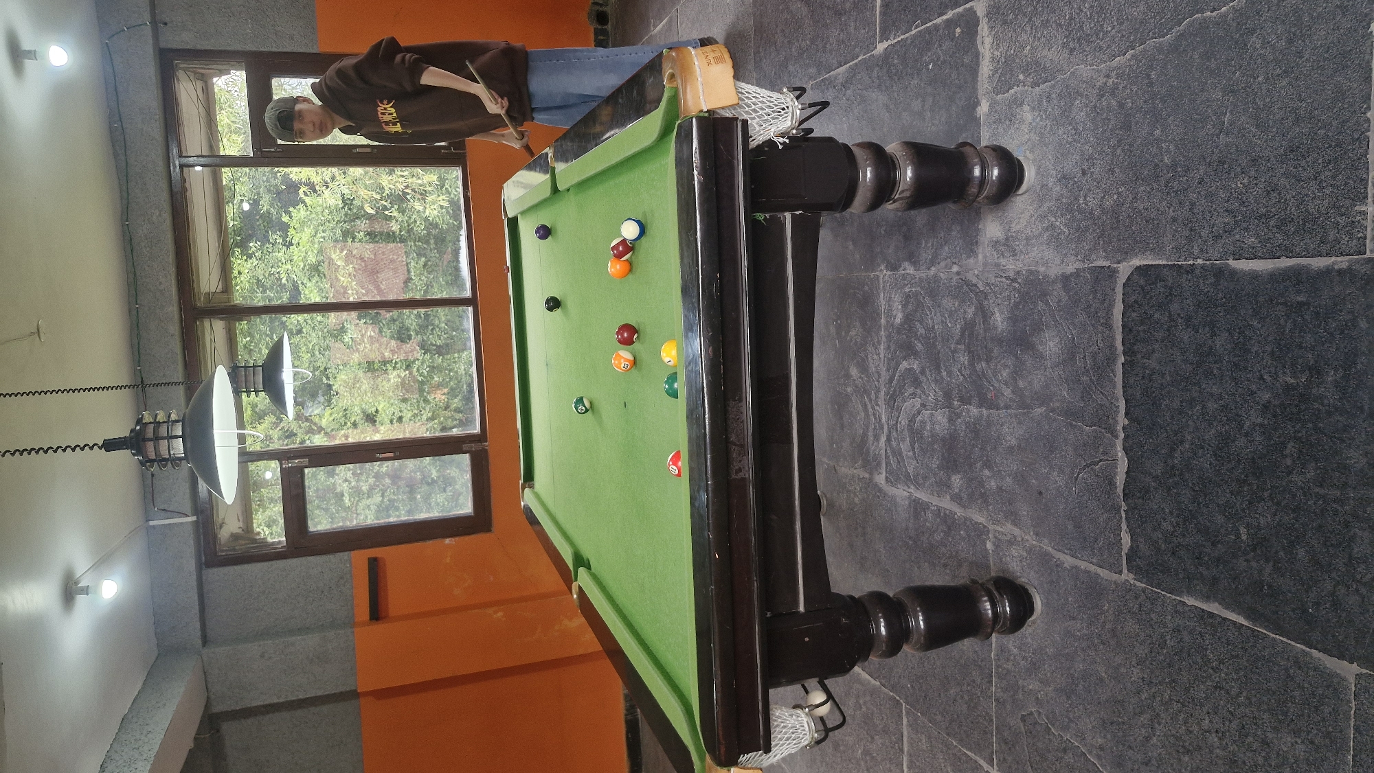 In Vashisht, India, a young person wearing a brown hoodie and a cap stands by a billiard table. They are holding a cue stick, preparing to strike a ball. Several billiard balls are scattered across the green felt surface of the table. The table is dark wood with ornate legs and is positioned in a room with orange walls and large windows that look out onto lush green trees. The floor is made of dark, irregular stone tiles. Two industrial-style pendant lights hang from the ceiling, illuminating the space.