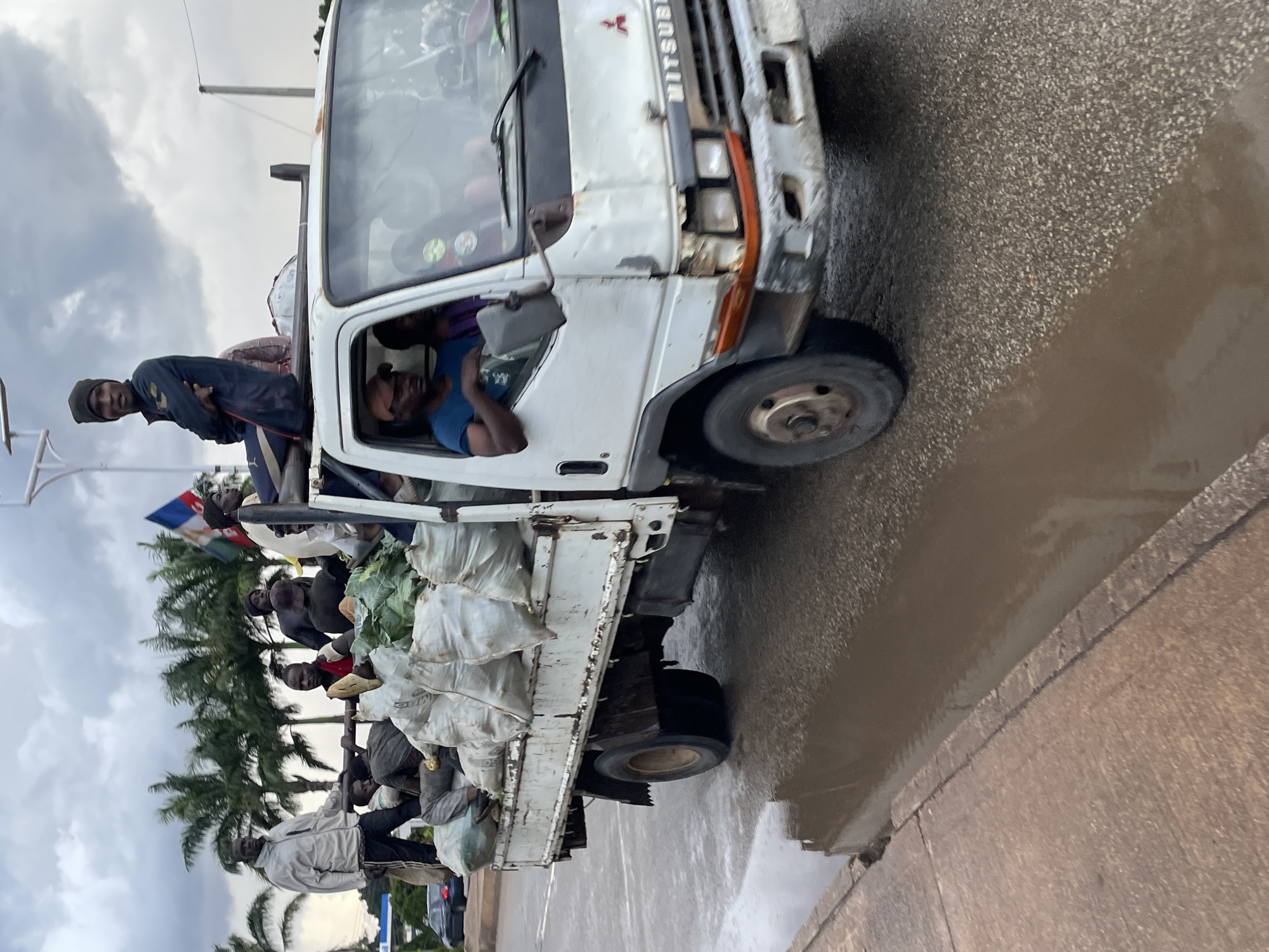 A flatbed truck is driving on a wet street, with a puddle of muddy water on the left side of the frame. The truck is laden with large, white sacks of produce and several people are riding in the back. A flag with red, white, and green stripes is visible. The truck cab is white and appears to be a Mitsubishi. Three men are visible in the cab. The overall impression is one of agricultural transport in a seemingly informal setting. The scene is located on a road in the city of Jos, Nigeria, where such informal transportation of goods and people is common.