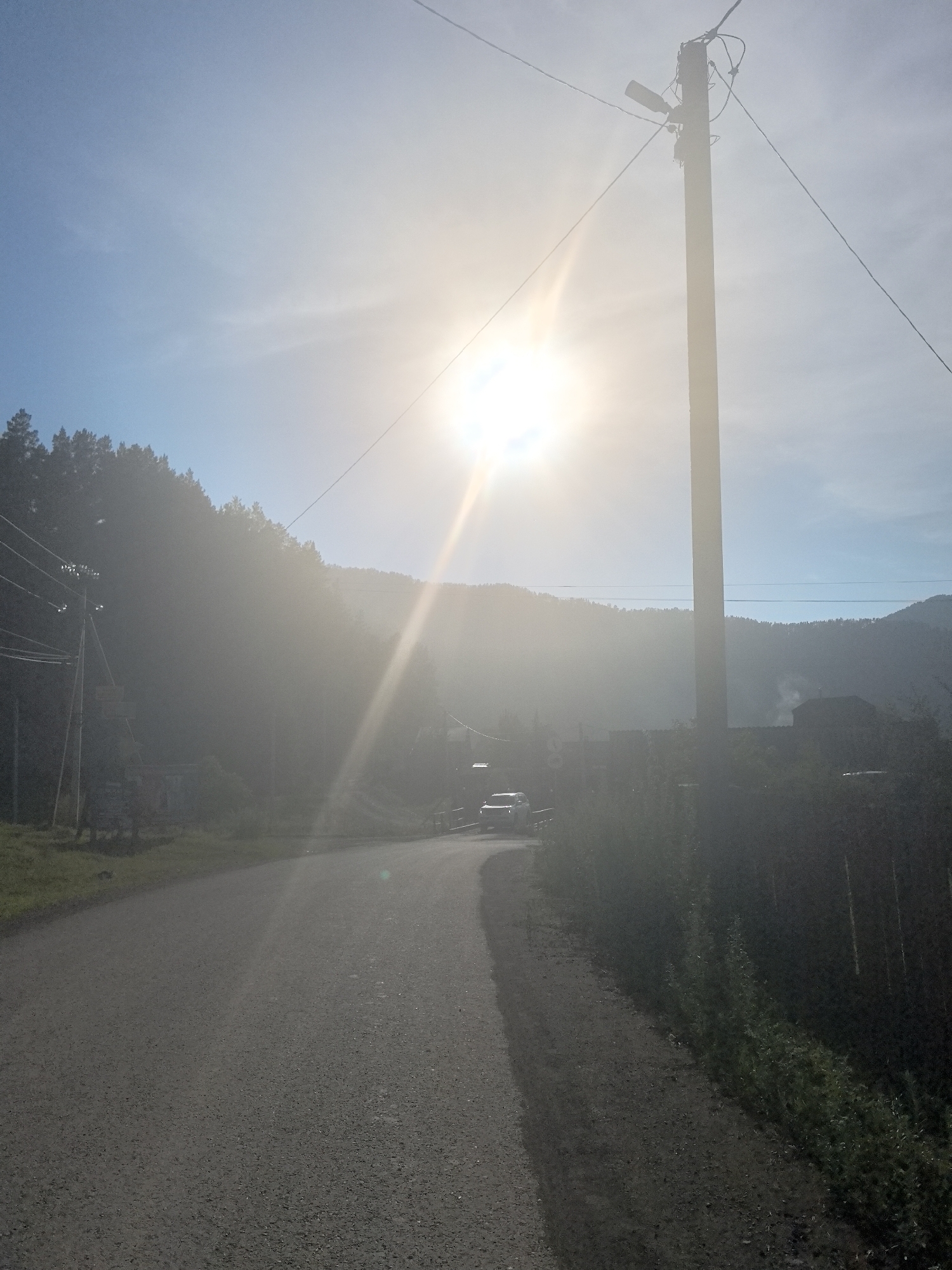 A bright sun, partially obscured by haze, shines down from a clear blue sky over a rural landscape. The sun is positioned in the upper left quadrant of the image, casting lens flare and creating a strong glare that washes out details in the distance. A tall wooden utility pole with wires and a light fixture stands prominently on the right side of the frame, silhouetted against the bright sky.
In the foreground, a paved road curves to the right, disappearing into the haze. The road appears to be asphalt with a gravel shoulder. To the left of the road, a dense evergreen forest covers a hillside, also partially obscured by the haze. On the right side of the road, a wooden fence runs along the edge of the property, with vegetation growing behind it.
Further down the road, a white vehicle is visible, appearing to be a truck or SUV, driving away from the viewer. In the background, hazy mountains form the horizon, with some scattered houses or structures visible among the trees. A faint plume of smoke rises from one of the structures in the distance. The overall atmosphere is hazy and sun-drenched, suggesting a warm day. The location is described as Unknown, Unknown.