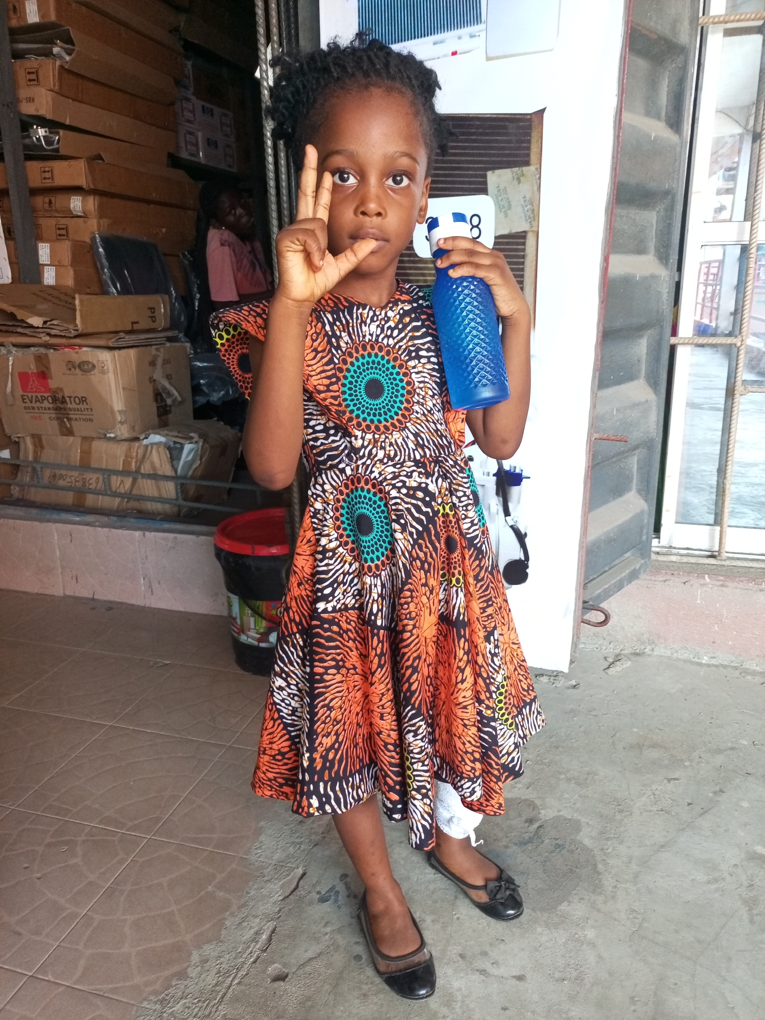 A young Black girl stands in what appears to be a storefront in Satellite Town, Nigeria. She is wearing a brightly colored African print dress, black flats, and holding a blue water bottle in her left hand. She is making a "V" sign with her right hand. Behind her are stacks of cardboard boxes and a person sitting in the background. The floor is tiled, and there is a doorway with a barred window nearby.