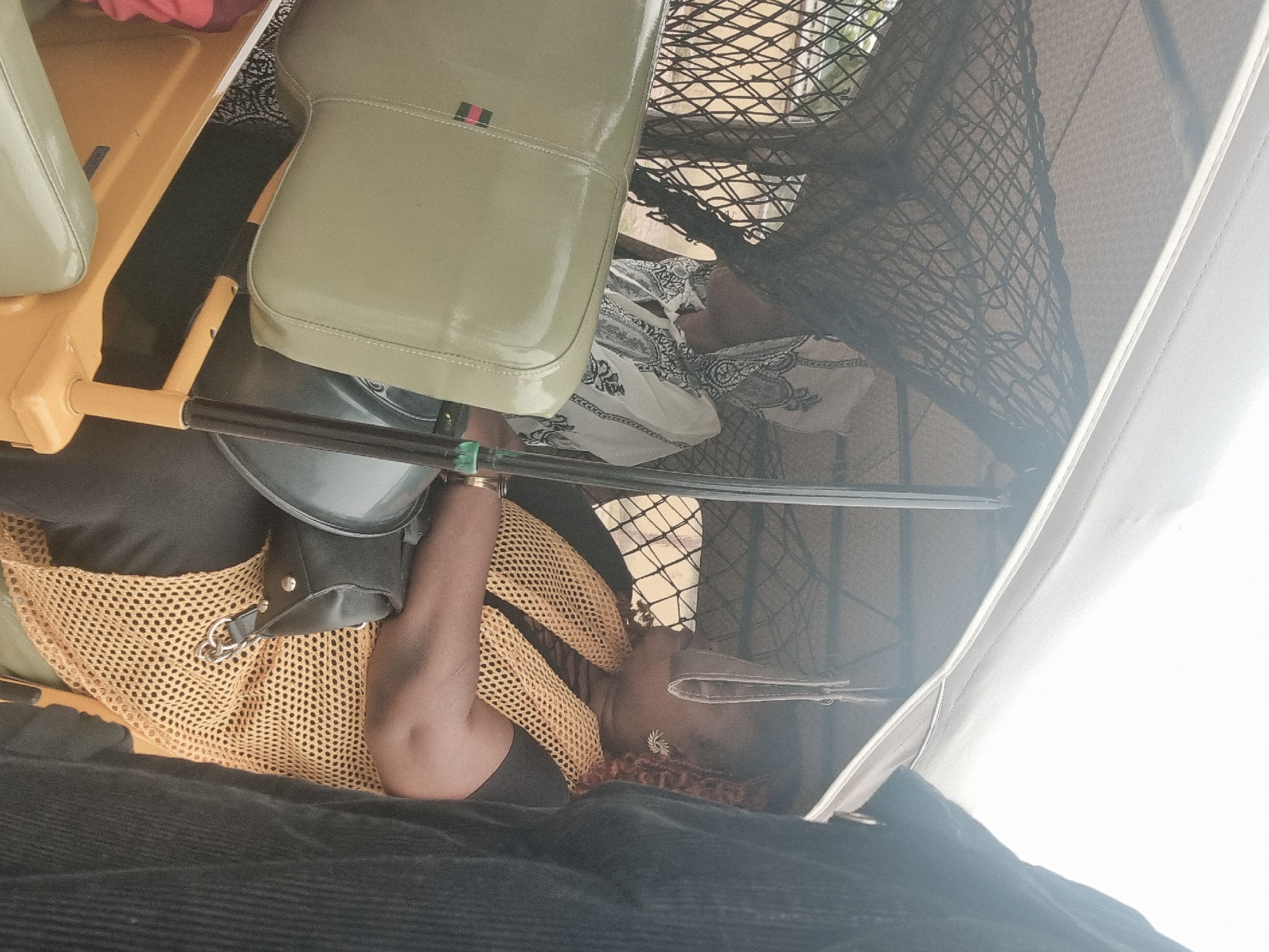 The image shows an interior view, potentially of a vehicle, with a person seated. The person is wearing a black top and a yellow, net-like garment. A bag with silver hardware is visible. Seating is present, including one seat that is a pale green color with stitching details. A net-like barrier is also visible. The location in the city of Jos, Nigeria, cannot be ascertained from the image.