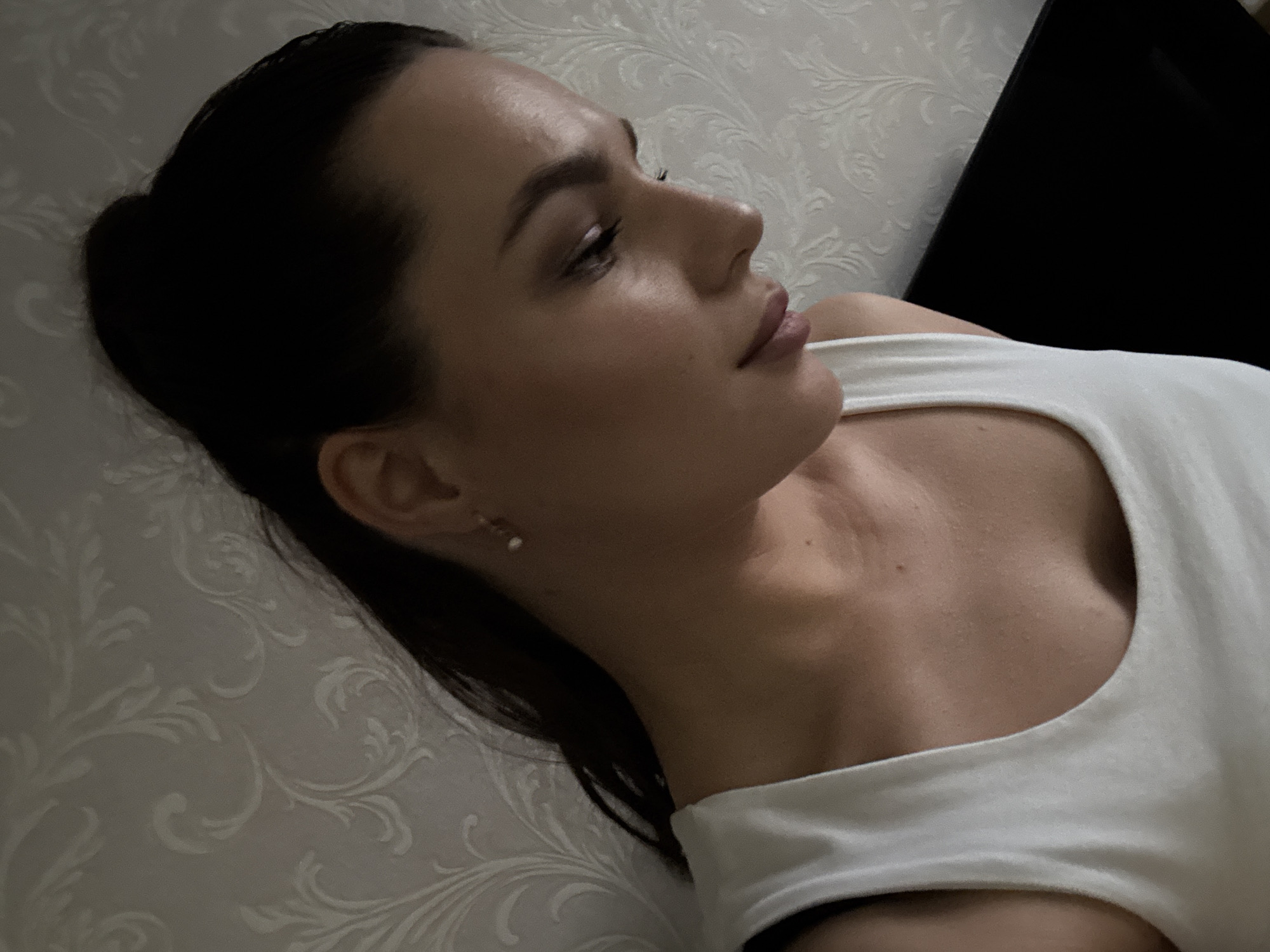 The image shows a close-up, over-the-shoulder shot of a young woman. She has dark hair pulled back into a ponytail and is wearing a white tank top. She is looking downwards and to her right, with her head tilted. Her right ear is visible, adorned with a small pearl earring. The background consists of a patterned wallpaper with an embossed floral design. The lighting is soft, creating a slightly moody atmosphere. A dark, flat surface, possibly a piece of furniture or a television, is visible in the lower right portion of the image. The scene appears to be indoors, in a residential setting. There is no indication of the specific location within the city of Chaikovsky, Russia.