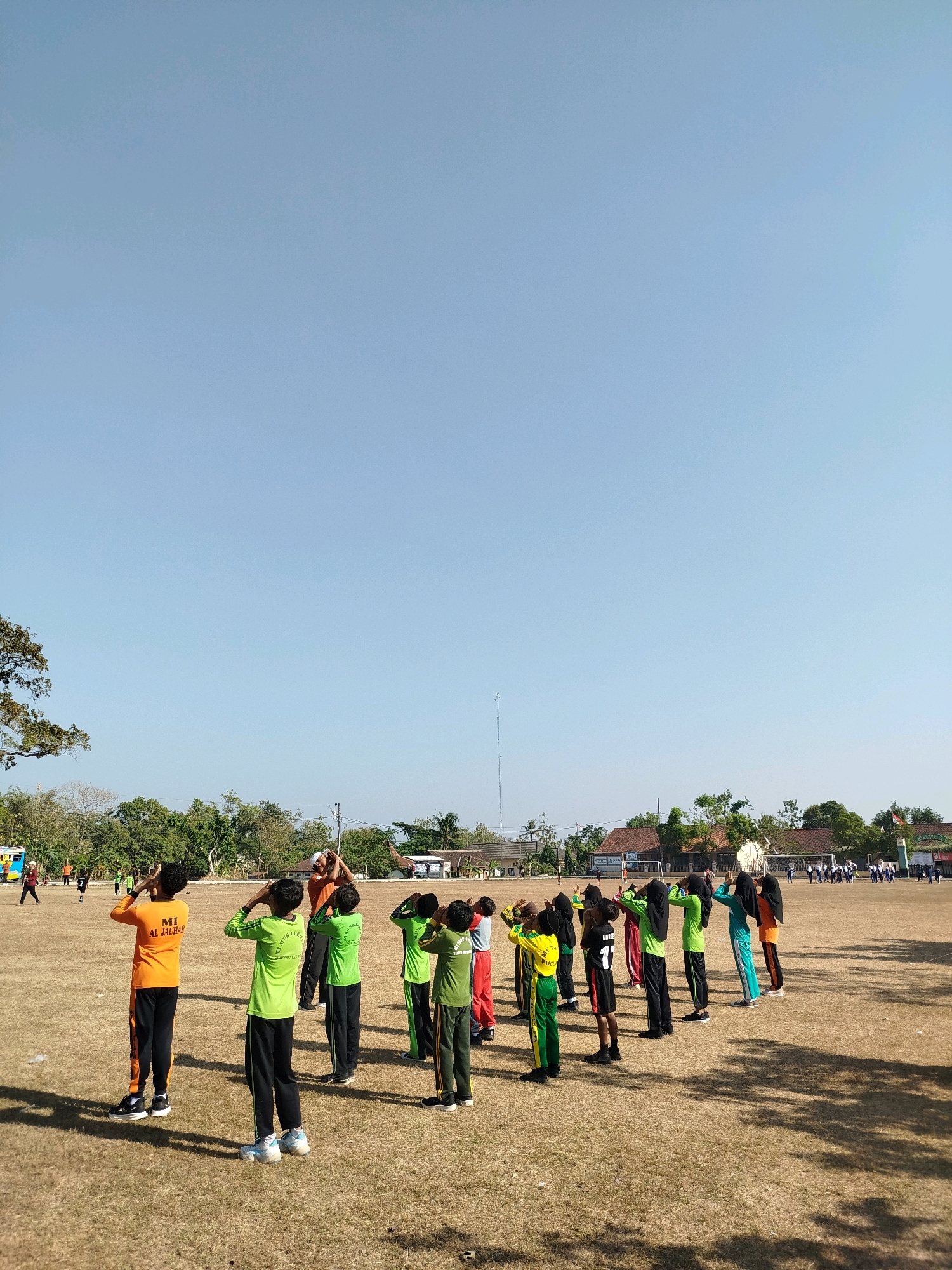 In Semin, Indonesia, a group of students are gathered on a dry, grassy field under a clear blue sky. The students, dressed in various brightly colored athletic attire, including orange, green, and black, are arranged in a line, facing away from the camera and performing some form of synchronized exercise or stretching. Their hands are raised to their heads, a common pose in warm-up routines. The field is sparsely populated with dry grass and patches of dirt. In the background, a line of buildings with reddish-brown roofs suggests a rural or suburban setting. Trees and vegetation are visible behind the buildings, and a tall radio tower stands in the distance. The overall scene depicts a group of students engaged in physical activity outdoors.