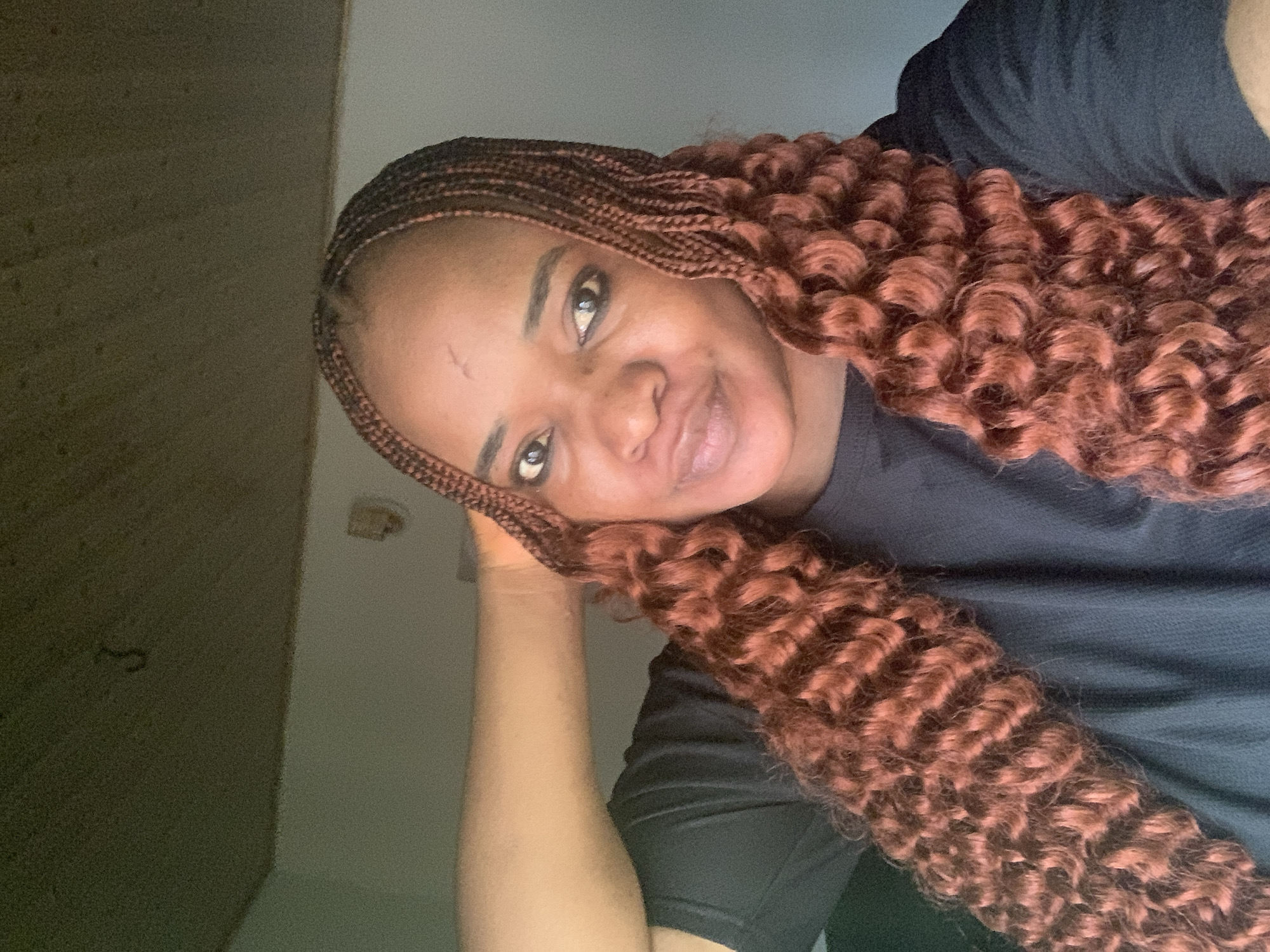 The image shows a woman with braided and curly reddish-brown hair, wearing a black shirt. She is leaning her head on her hand, and looking at the camera with a slight smile. The background is a plain wall with a light fixture or switch. There is no indication of the location being in Barakin Akawo, Nigeria. The location is generic.