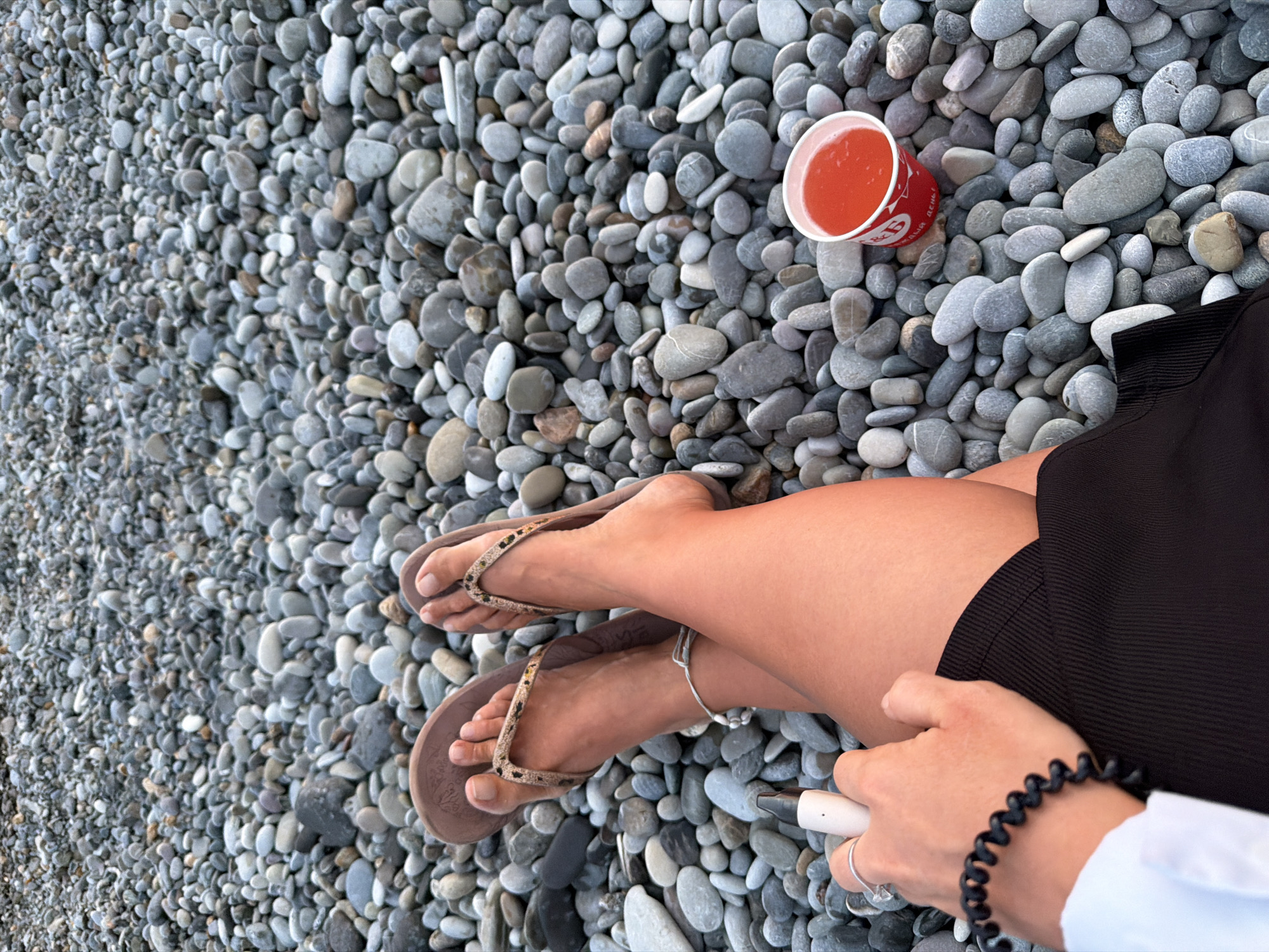 A person sits on a beach covered in smooth gray and white pebbles. Their legs are crossed, wearing brown flip-flops with a leopard print strap and a black skirt. They hold a white cylindrical object in their right hand, with a ring on their index finger and a black spiral bracelet on their wrist. To their right, a red and white paper cup filled with a pink beverage sits on the pebbles. The background consists entirely of the pebble-covered beach.