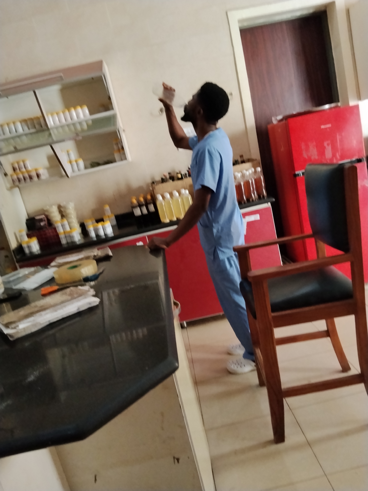 A person in a blue medical scrub suit is standing in a laboratory or pharmacy in Gwarinpa, Nigeria. The individual is holding a clear plastic cup to their mouth, seemingly drinking. The room contains shelves stocked with numerous bottles and containers, a red refrigerator, a dark countertop, and a wooden chair with a dark upholstered seat. The environment appears to be a functional space with various medical or pharmaceutical supplies.