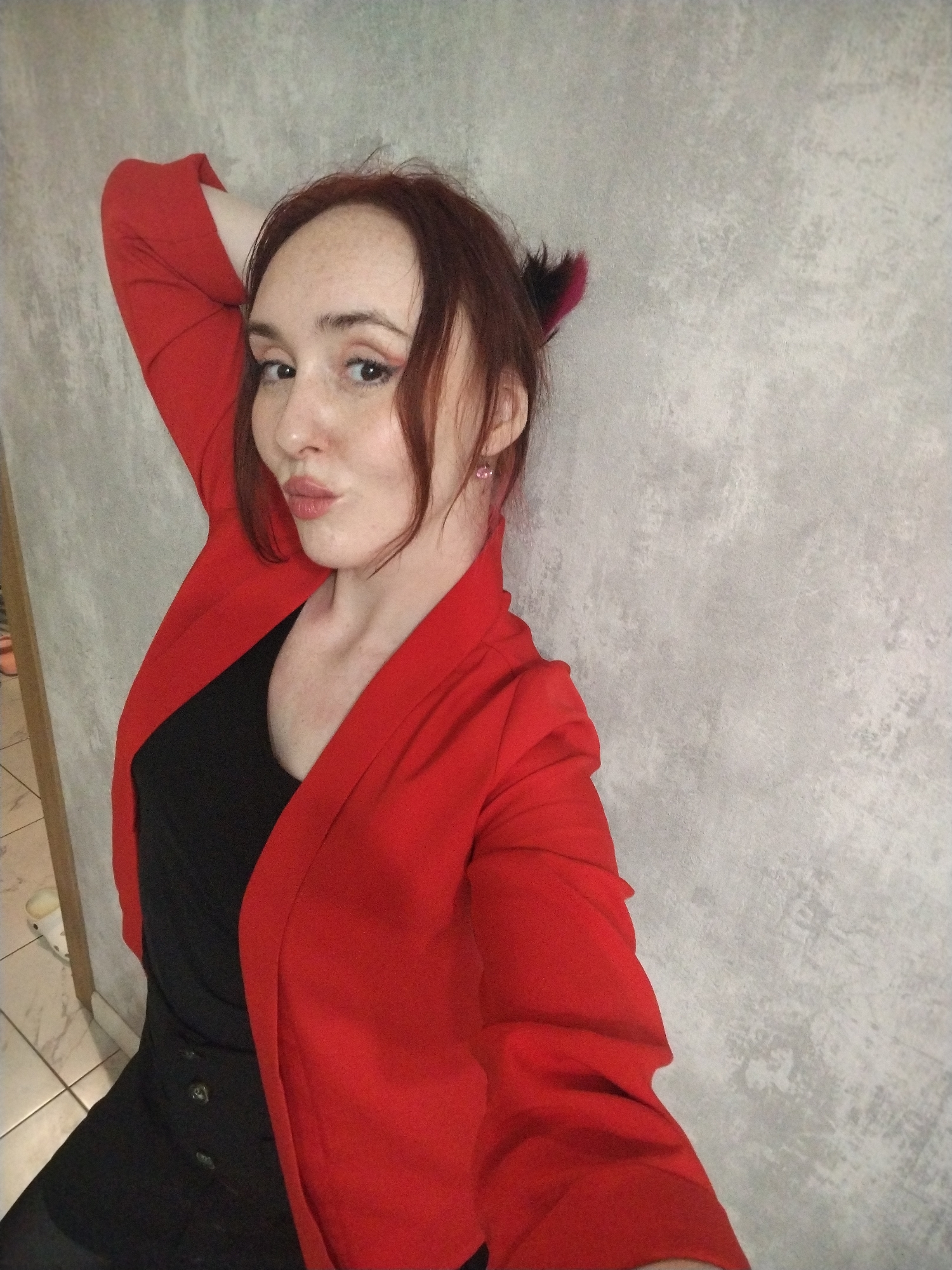 A woman stands against a textured gray wall. She is wearing a red blazer over a black top and bottom. Her left arm is raised and bent behind her head. Her hair is reddish-brown, and she has a small, decorative item with black and red accents in her hair near her right ear. The location is not possible to determine from the image.