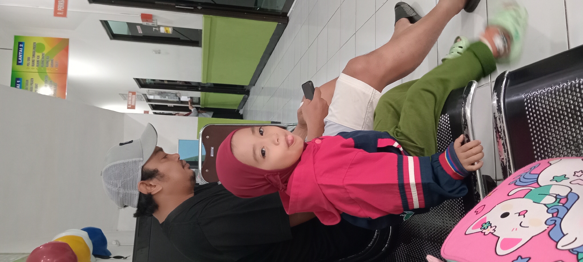 In an indoor waiting area, likely a clinic or hospital in Mataram, Indonesia, a man wearing a gray mesh baseball cap and a black t-shirt sits to the left. He is looking down, holding a dark smartphone. Beside him, a young child with a red hijab and a pink and blue jacket sits on a black chair, sticking their tongue out playfully. The child wears green pants and light green sandals, with their right foot in motion. In the foreground to the left, a pink bag with a cartoon cat design is visible on another black chair. The environment features a tiled floor and a hallway with several doors and green wall accents. Overhead signs indicate directional information, one reading "AI 1" and "LANTAI 2" which translates to "Floor 2". Another sign partially visible reads "R. INSTALASI FARMASI" which translates to "Pharmacy Installation Room".
