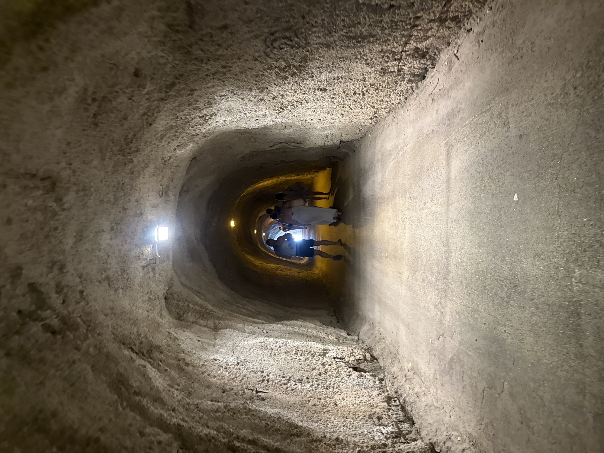 The image depicts an underground tunnel with rough, textured walls. The perspective is from within the tunnel, looking towards a group of people at the far end. The tunnel appears to be illuminated by artificial lights, with one visible light fixture on the ceiling to the left, and softer light emanating from the distance where the people are. The floor of the tunnel is dirt or gravel, and the walls are uneven, suggesting it may be a natural formation or an ancient excavation.
There are at least four people visible in the distance, appearing to walk or stand within the tunnel. They are mostly silhouetted against the light, making it difficult to discern details about their clothing or appearance, though some are wearing lighter colored garments. One person is seen lying on their back on the tunnel floor, with their head towards the opening and their feet towards the far end. The overall atmosphere of the scene is somewhat mysterious due to the dim lighting and enclosed space.
The texture of the tunnel walls varies, with some areas appearing smoother and others rougher and more porous. The color palette is dominated by earthy tones of beige, brown, and gray. There are no visible signs or text within the image. The scene could be interpreted as a tour group exploring an underground site. The lack of natural light and the enclosed nature of the tunnel suggest an indoor or subterranean environment, consistent with the provided location context of Kamenovo, Montenegro, which is known for its caves and historical sites.
