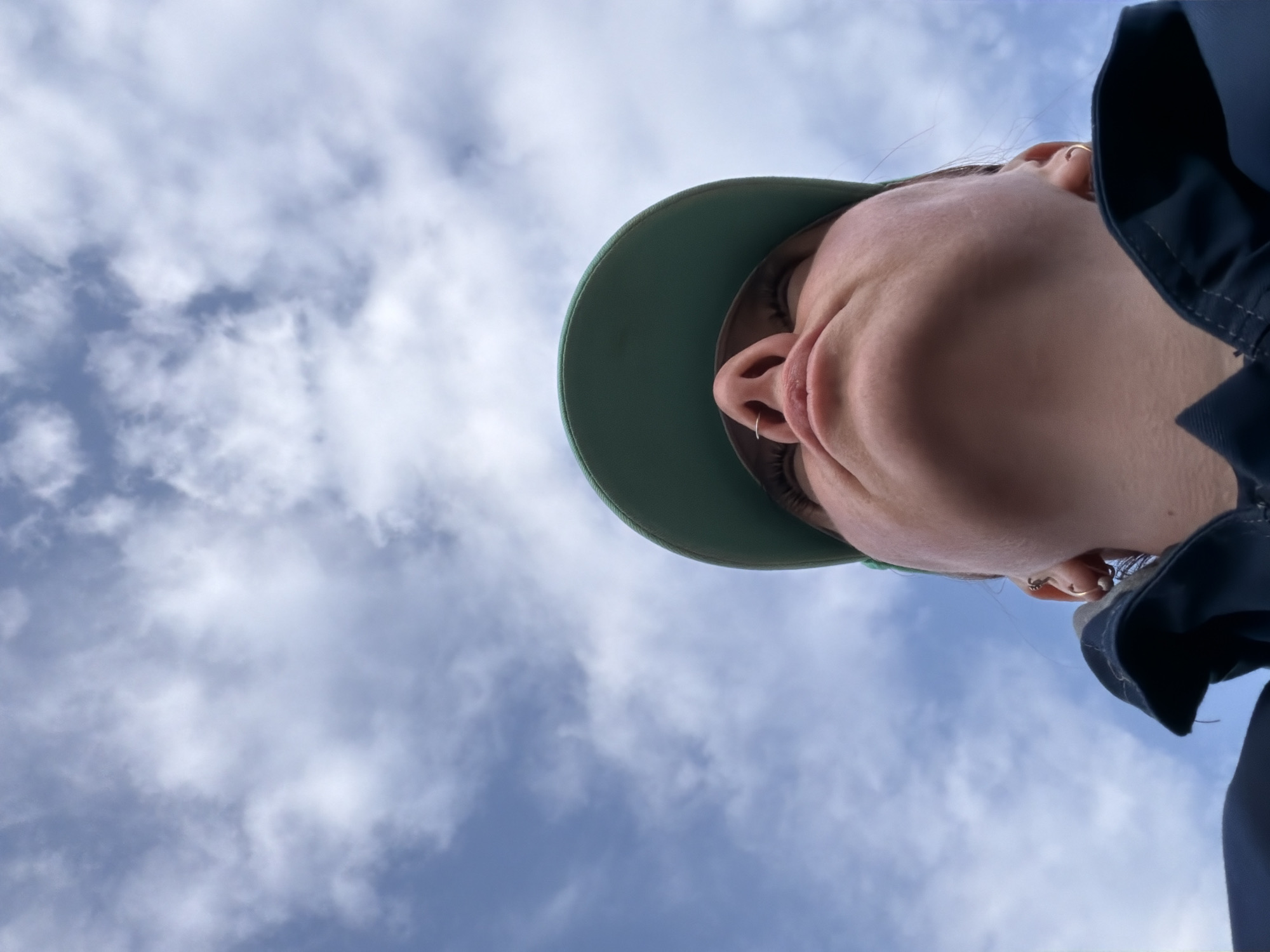 A low-angle shot features a person wearing a green cap and a dark blue jacket, with a cloudy blue sky in the background. The person's face, specifically the nose, mouth, and chin, are prominently visible. The subject's earlobes are pierced with gold hoops. The location is indiscernible based on the image.