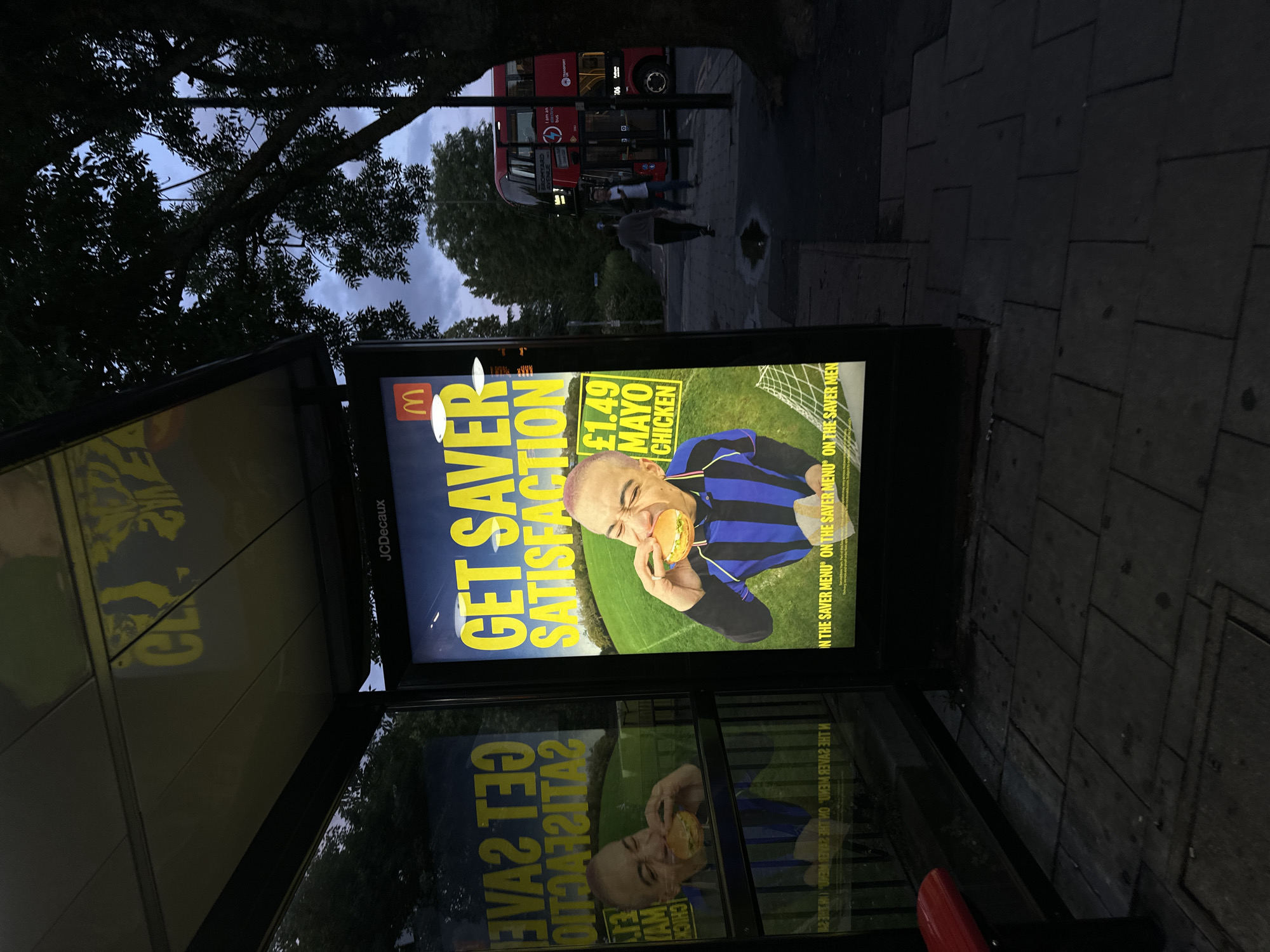 The image captures a bus stop scene at dusk or night, indicated by the dark, overcast sky and the illumination of the advertisement within the bus shelter. The primary subject is a brightly lit McDonald's advertisement poster displayed inside the bus stop.
The advertisement features a person, a man with short, light-colored hair, enthusiastically biting into a burger. He is wearing a blue and black striped jersey, reminiscent of a football (soccer) kit, and is positioned on what appears to be a green sports field with a goal net in the background. The text on the ad reads "GET SAVER SATISFACTION" prominently in large yellow letters. Below this, it states "£1.49 MAYO CHICKEN". The McDonald's 'M' logo is visible in the top left corner. Smaller text, partially repeated, says "IN THE SAVER MENU".
The ad is reflected in the glass panel of the bus stop, creating a mirrored and slightly distorted image below the main display. The bus stop shelter itself has "JCDecaux" branding visible on its side, a common street furniture provider in urban areas.
Beyond the bus stop, a red double-decker bus, a hallmark of London transport, is visible on the street. Its route number, "106", is discernible, along with a partially visible sign that reads "I am an bus". A few indistinct figures of people can be seen near the bus on the paved sidewalk. Silhouetted tree branches frame the top of the image against the dark sky, adding to the nighttime atmosphere. The ground around the bus stop is paved with dark grey square slabs.