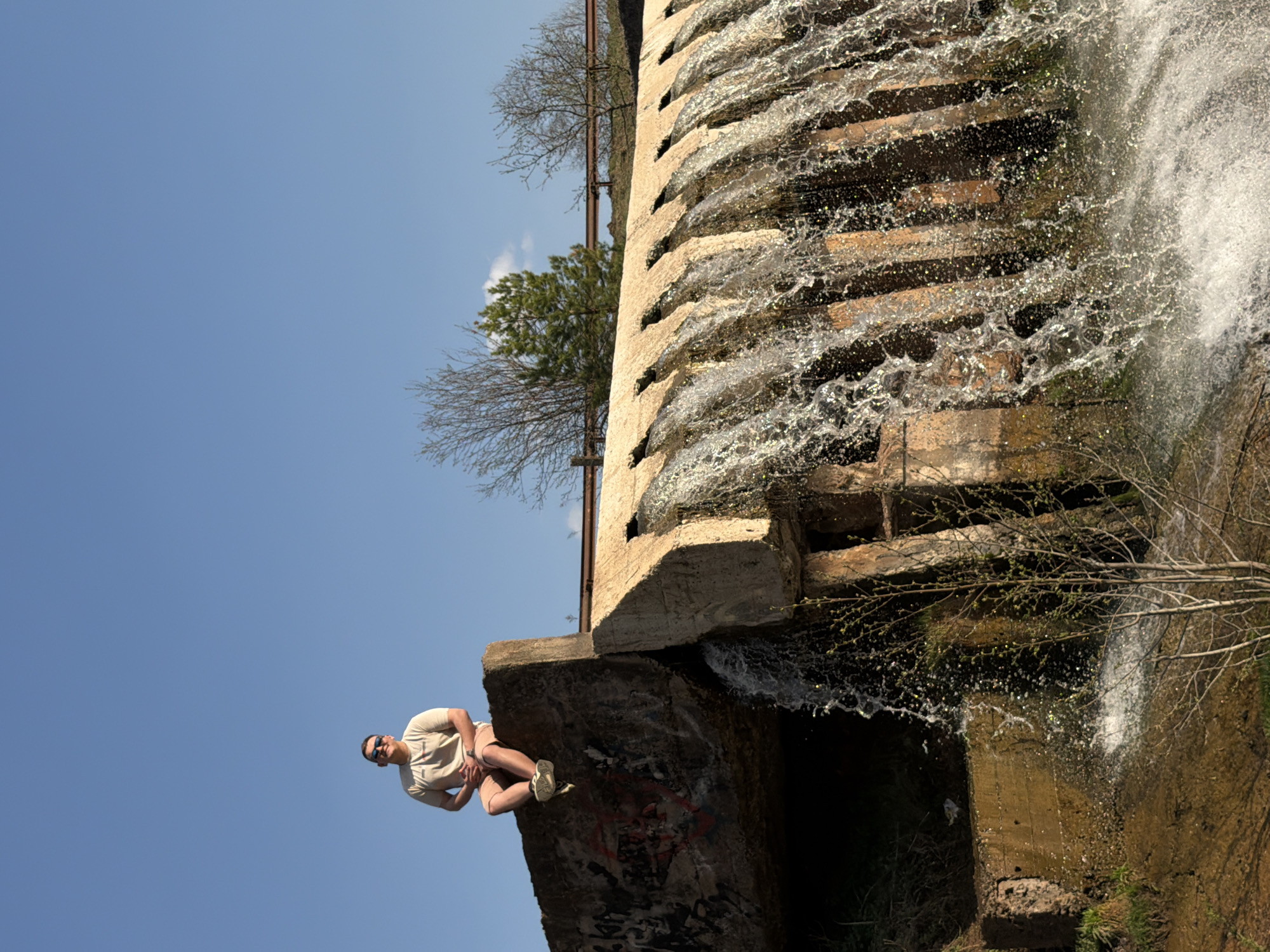 The image depicts a man sitting on a concrete ledge adjacent to a man-made waterfall structure in Kamennoye, Russia. The man is wearing a light-colored t-shirt, shorts, and sunglasses, and is facing the camera. The waterfall consists of multiple tiers of concrete blocks, with water cascading over each level. The surrounding environment includes bare trees and vegetation. The sky is clear and blue.