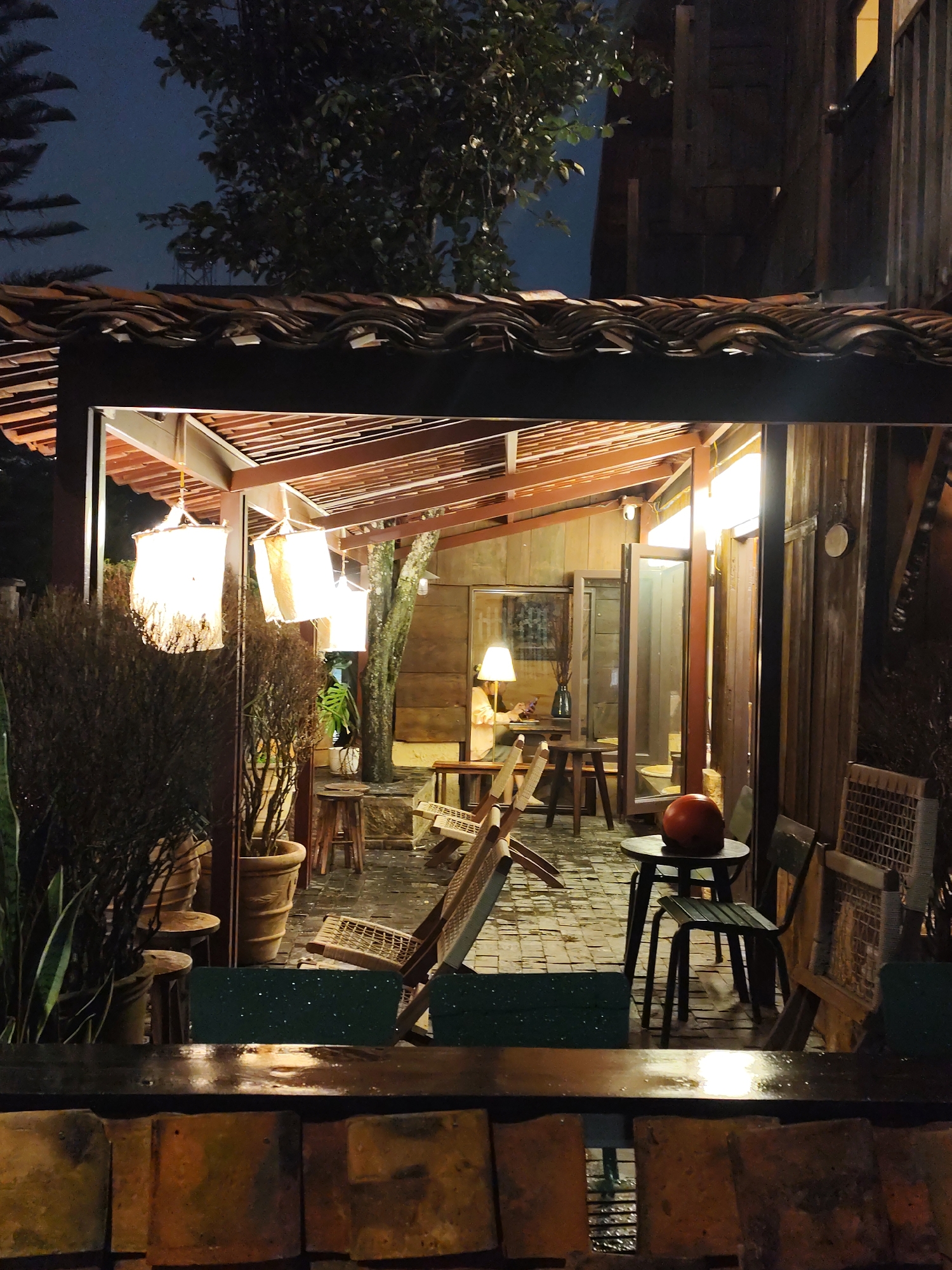 The image depicts an outdoor seating area of a cafe or restaurant in Da Lat, Vietnam, at night. The scene is illuminated by warm, artificial lighting emanating from hanging lanterns and lamps within the covered structure. The architecture features a tiled roof supported by wooden beams, with walls made of wood and glass doors.
In the foreground, a low wooden structure with textured tiles is visible, possibly a bar or counter, with reflections on its wet surface. Beyond this, several chairs and tables are arranged in the seating area. To the left, there are large potted plants with dense, bushy foliage and a tall, thin plant.
Within the covered area, several woven lanterns hang from the ceiling, casting a soft glow. One lamp with a white shade sits on a table inside, illuminating a section of the space. A tree with a visible trunk and branches grows within the covered area, adding a natural element.
Further inside, a person is seated at a table, looking at their phone. Open glass doors reveal more of the interior, which appears to be decorated with wooden paneling and possibly artwork on the walls. Various chairs, some made of wood and woven material, are scattered throughout the seating area. On a small round table in the middle ground, an orange, round object, possibly a decorative pumpkin or gourd, is placed. The ground surface appears to be paved with cobblestones or similar material and is wet, suggesting recent rain.
The overall ambiance is cozy and inviting, with the dim lighting and natural materials creating a rustic yet sophisticated atmosphere. The presence of lush greenery and the architectural style are consistent with the cool climate and hilly terrain of Da Lat.