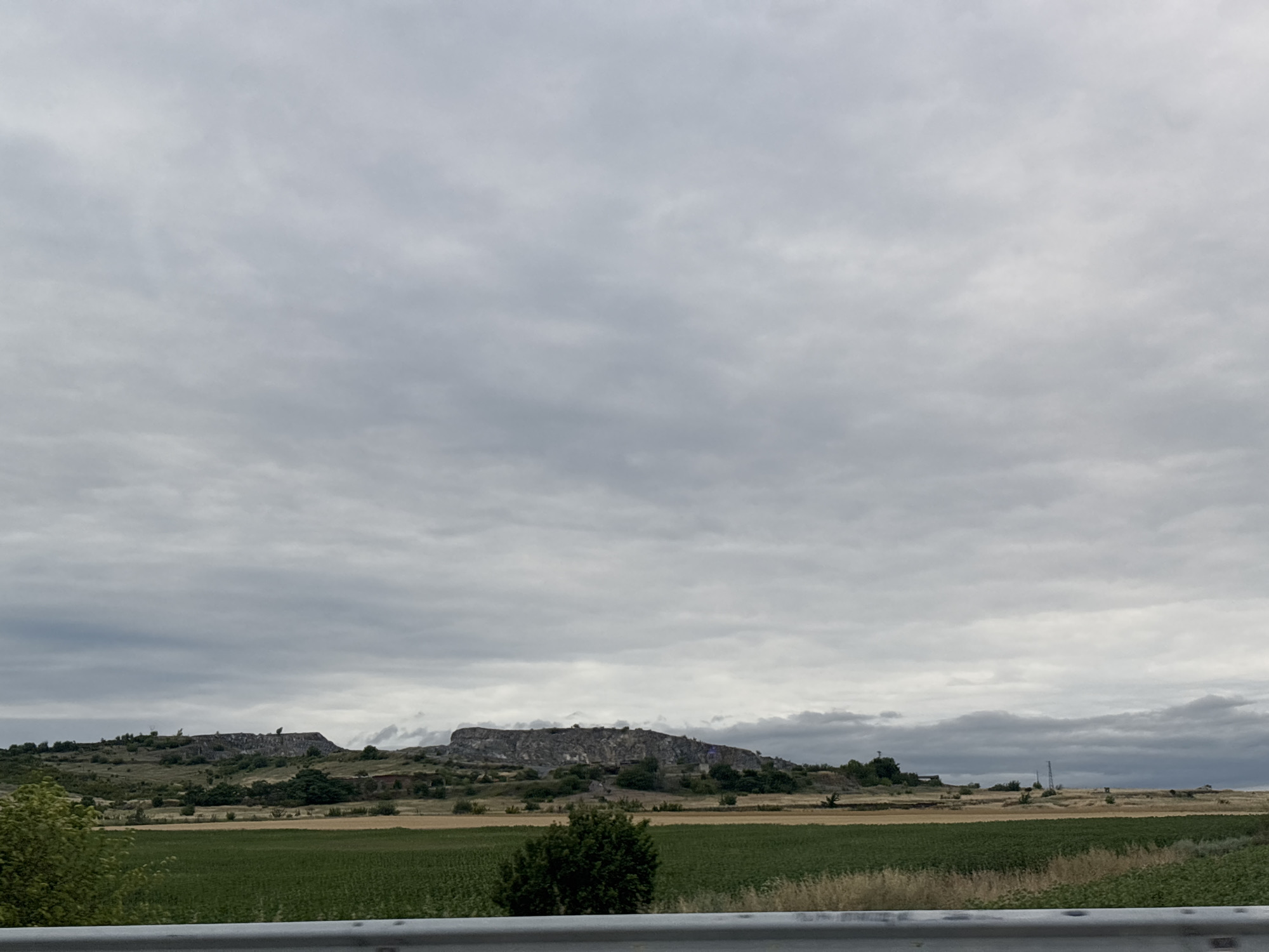 A wide shot captures a desolate, overcast landscape with a large, rocky hill formation dominating the middle ground. The hill is covered in vegetation and shows signs of human activity, possibly quarrying or ancient structures. In the foreground, a cultivated field of green crops is visible, separated by a rough, grey barrier in the lower part of the frame. The sky is filled with heavy, grey clouds, suggesting a gloomy or impending weather event. The scene appears to be a rural area, potentially near Polsko Padarevo, Bulgaria, given the rocky terrain and agricultural fields. There are no people or animals visible in the image, and the overall impression is one of natural ruggedness mixed with subtle signs of human impact.