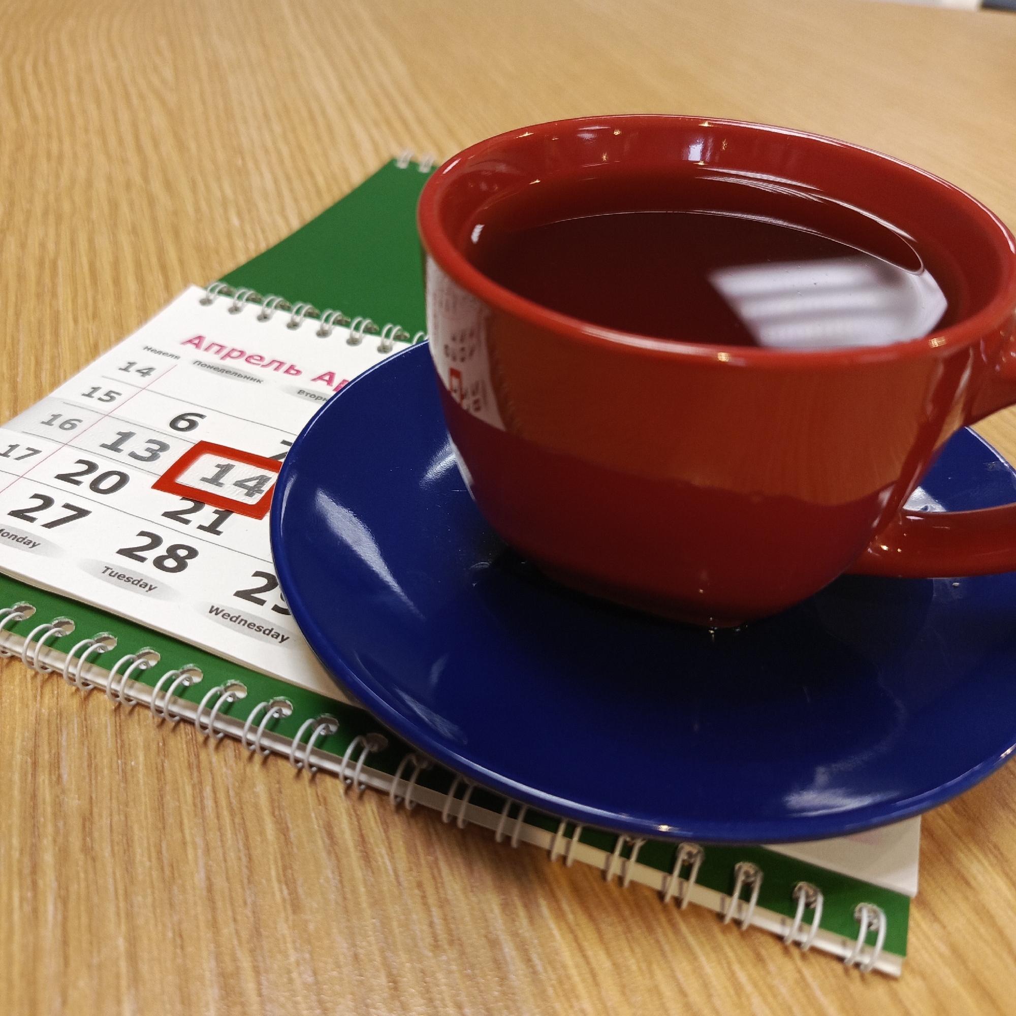 The image depicts a red cup filled with a dark liquid, resting on a blue saucer. The saucer is placed on top of a desk calendar with a green cover. The calendar is open to the month of April, and the date "14" is highlighted in red. The calendar pages are secured with a silver metal spiral binding. The surface the calendar and cup rest upon appears to be a light-colored wooden desk. The calendar has some text in Russian, and it is highly probable that the image was captured in a location in Russia. The calendar is open to April, the text "Апрель" indicates that the scene is related to the city of Chaikovsky, Russia.