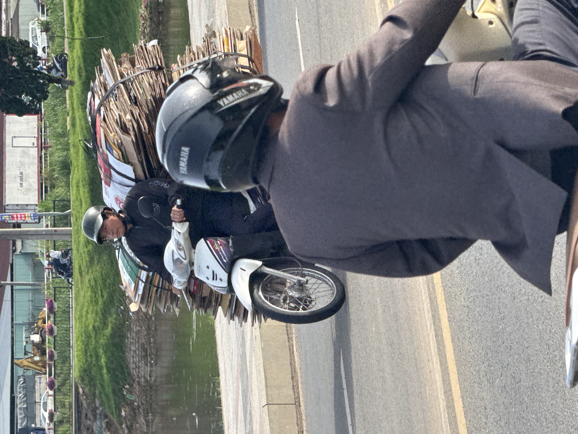 A motorcycle carries a large load of cardboard on a street in Da Lat, Vietnam. The rider, wearing a dark helmet and jacket, is positioned on the left side of the frame, facing right. The motorcycle itself is white and black. Behind the first rider, another person on a motorcycle, wearing a dark suit and a black helmet with "YAMAHA" visible, is seen from the back, facing away from the camera. They are both on a paved road with a yellow line. In the background, a body of water, a grassy embankment, a sidewalk, and various buildings are visible. A yellow excavator is present in the far background, suggesting a construction or industrial area. The sky is overcast.