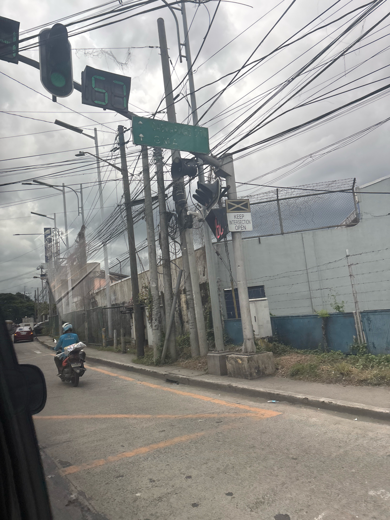 The image captures a street scene in Carmona, Philippines, under an overcast sky. A traffic light with a green signal is visible in the upper left, accompanied by a digital countdown timer displaying "59". Further down the street, another traffic light is visible, and a sign indicates "Keep Intersection Open". A motorcycle rider, wearing a blue shirt with "ANIKAS" printed on it and a white helmet, is proceeding away from the camera. The road is asphalt with yellow intersection markings. A red car is parked on the left side of the road. The background features a concrete wall with a chain-link fence on top, and numerous electrical wires crisscross the sky.