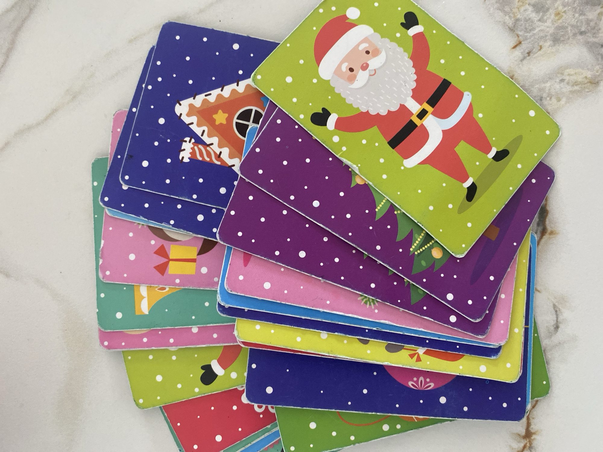 A collection of approximately 15 to 20 brightly colored, Christmas-themed playing cards or flashcards is fanned out and overlapping on a light-colored surface with a subtle, gray-brown marble pattern. The cards feature various holiday illustrations against backgrounds of green, red, pink, blue, purple, yellow, and teal, all uniformly adorned with white dots resembling falling snow. Specific illustrations include a smiling Santa Claus in a red suit on a green card, a gingerbread house with white frosting details and a star on a blue card, a decorated green Christmas tree on a purple card, and a yellow gift box with a red bow on a pink card. The cards exhibit minor wear and chipping along their edges. The scene is presented from a slightly elevated, overhead perspective. No specific geographical features or landmarks are discernible in the image.