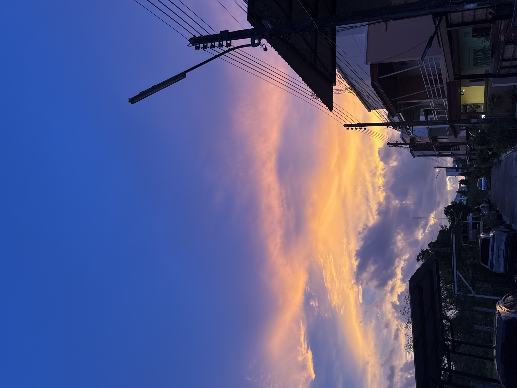 A street scene in Karon, Thailand, captures a vibrant sunset. The sky is a gradient of deep blue at the top, transitioning to hues of orange and pink where clouds are illuminated by the setting sun. A prominent power pole with multiple wires and a streetlight stands on the right side of the image, silhouetted against the colorful sky. To the right of the pole, a multi-story building with balconies and a sloping roof is visible. On the left side, another structure with a dark, corrugated roof is partially visible, with cars parked underneath. A paved road runs through the center of the image, with several cars parked along its side. The environment suggests a residential or semi-urban area.