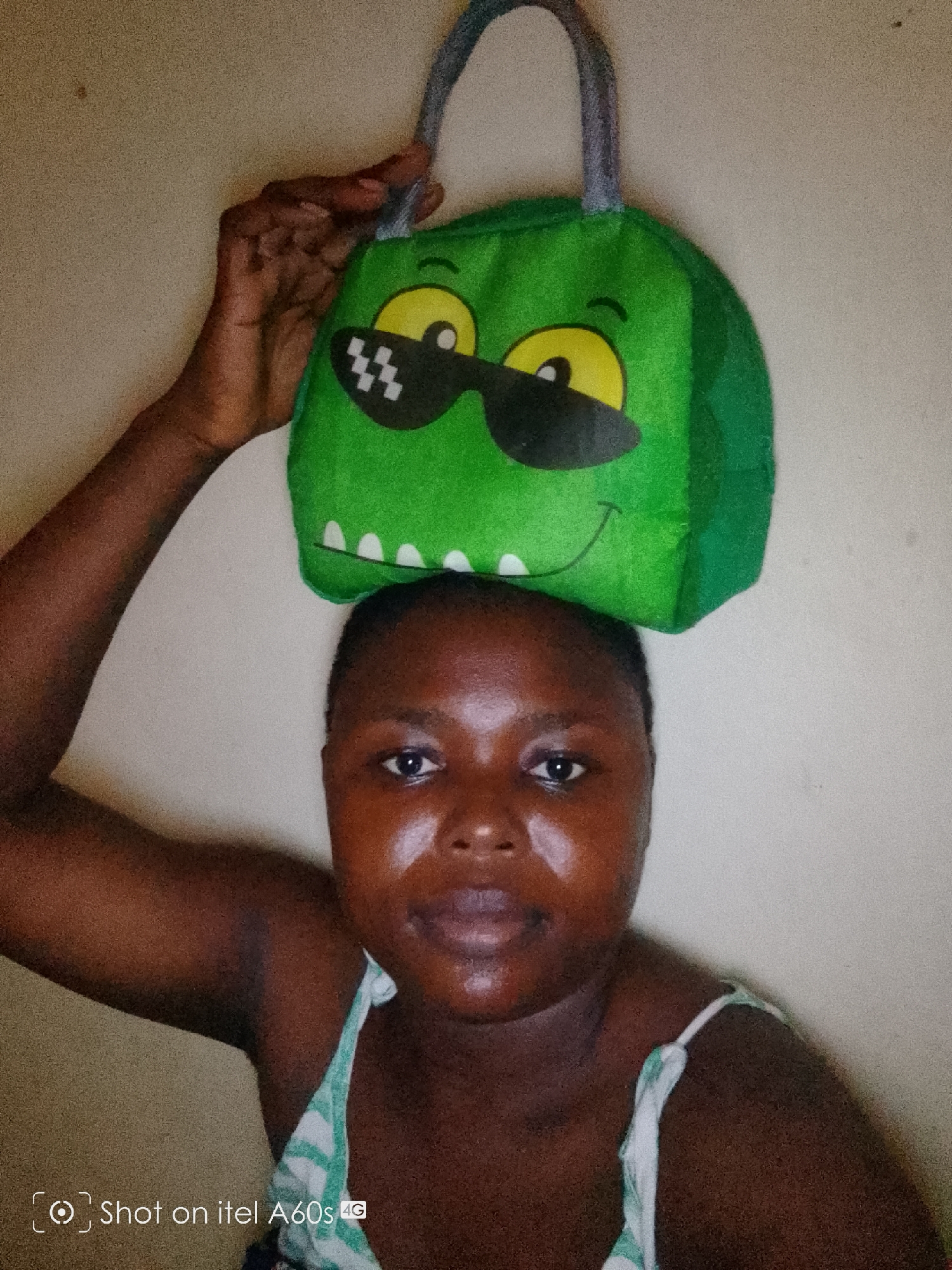 A woman with dark skin is holding a green bag with a cartoon face on her head. The woman is wearing a white and green striped top. The background is a plain, light-colored wall. The image was taken with an Itel A60s 4G phone. The location is Unknown, Unknown.