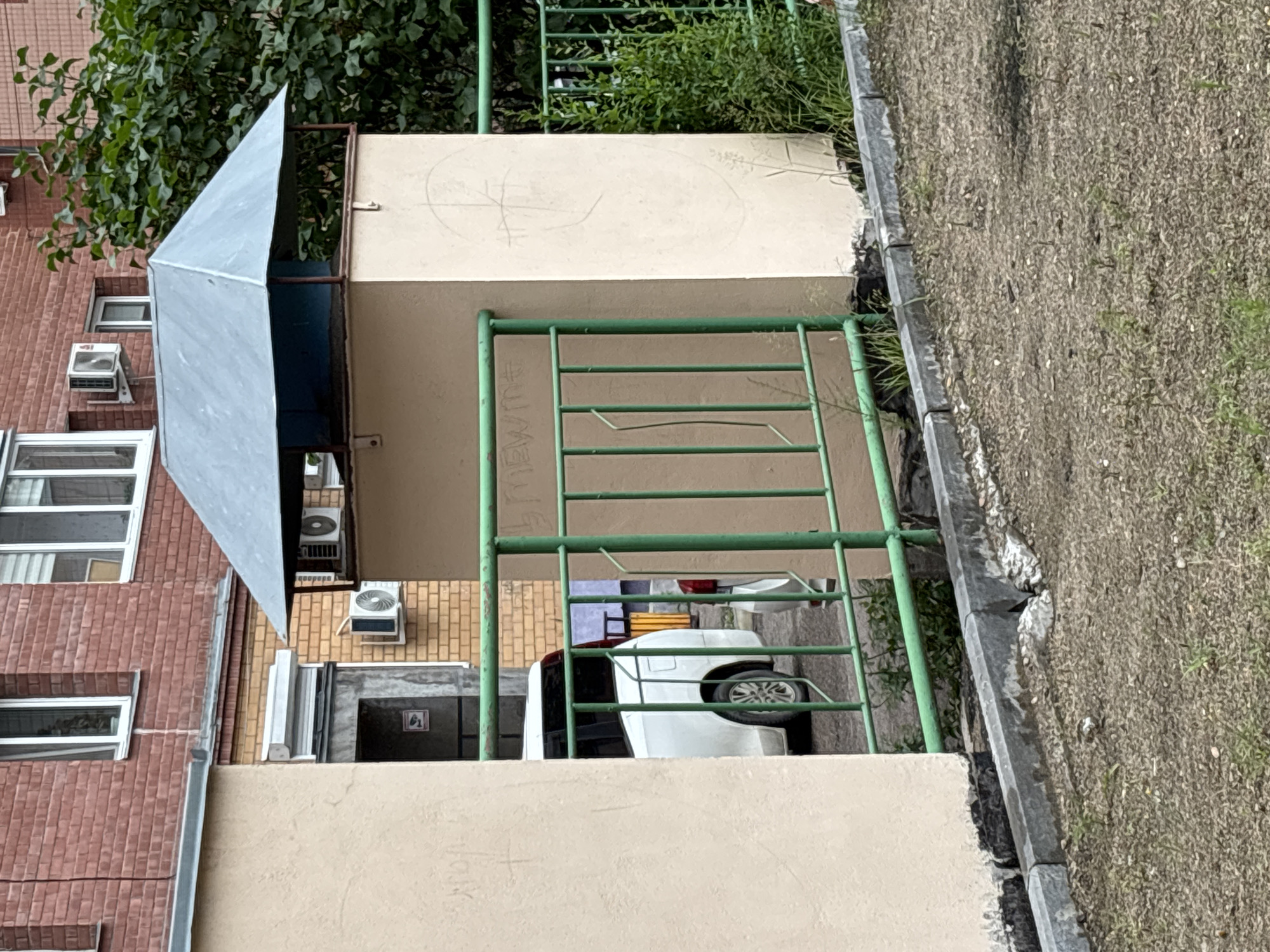 This media file shows an outdoor scene in Blagoveshchensk, Russia. The image is oriented vertically, with the bottom of the frame appearing at the top and vice versa. In the foreground, there is a light-colored wall with some faint graffiti. Behind this wall, a green metal railing encloses an area. Within this enclosed space, a white car is parked, partially obscured by the railing.
Beyond the car and railing, elements of a building are visible. To the left, a brick building with multiple windows and what appear to be air conditioning units is present. To the right of the brick building, a lighter-colored structure with a metallic, angled roof is seen. This structure is adjacent to another building facade made of yellowish brick, also featuring air conditioning units. Further back, behind the green railing, there are trees and other greenery.
The image appears to be taken during the daytime, with overcast or diffused lighting, suggesting a cloudy or possibly rainy day. The setting suggests an urban or suburban environment, possibly a courtyard or residential area. No people are visible in the image.
Notable details include the graffiti on the wall and the metal railing, which seems to be part of a playground structure or a protective barrier. The car is a white sedan. The overall impression is that of a somewhat utilitarian and unkempt outdoor space.