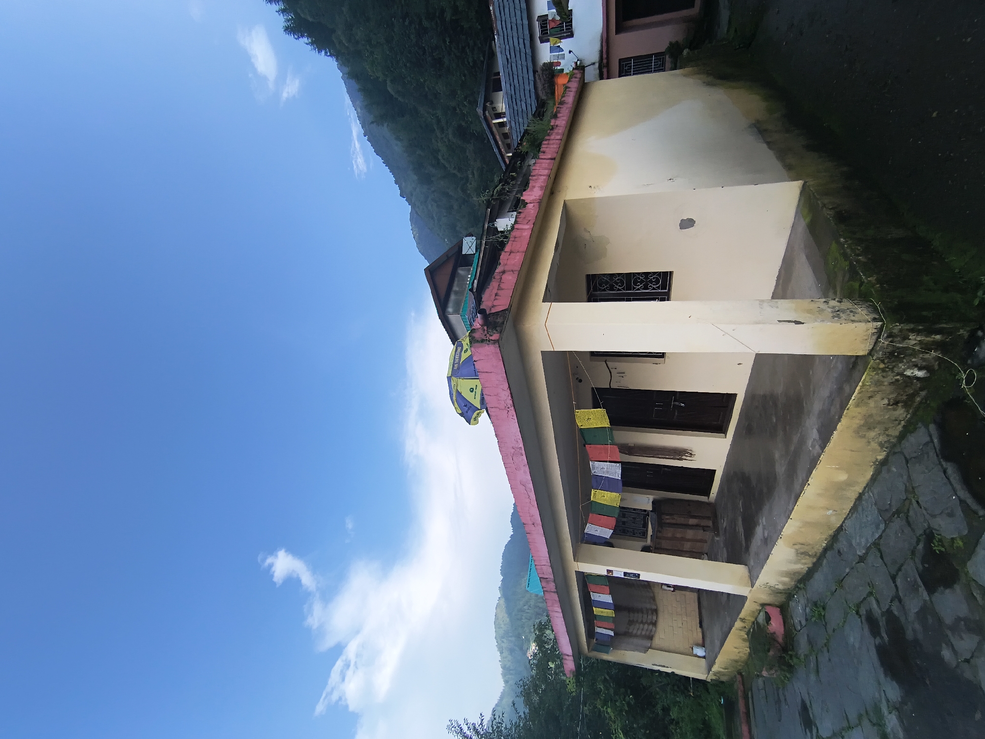 The image shows a building in Haini, India, with a clear blue sky and some clouds above. The building is light-colored with a pink trim along the roof. Prayer flags are strung across the front of the building. In front of the building is a paved area that appears to be wet, suggesting recent rain. To the right and behind the building, there are more buildings and a lush green hillside covered in trees. There are no people visible in the image. The overall scene depicts a quiet, possibly rural or semi-rural, setting in a mountainous region.