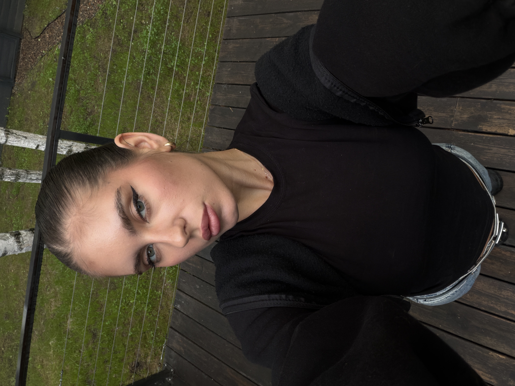 A woman is captured in a selfie from a high angle, looking directly at the camera. She has her hair pulled back in a tight bun and is wearing makeup, including eyeliner and lipstick. She is dressed in a black long-sleeved shirt and jeans. The background consists of a wooden deck and lush green grass, with birch trees visible in the distance. The scene suggests an outdoor setting, possibly a yard or a garden area. The provided location information states the image was taken in the city of Berezovka, Russia.