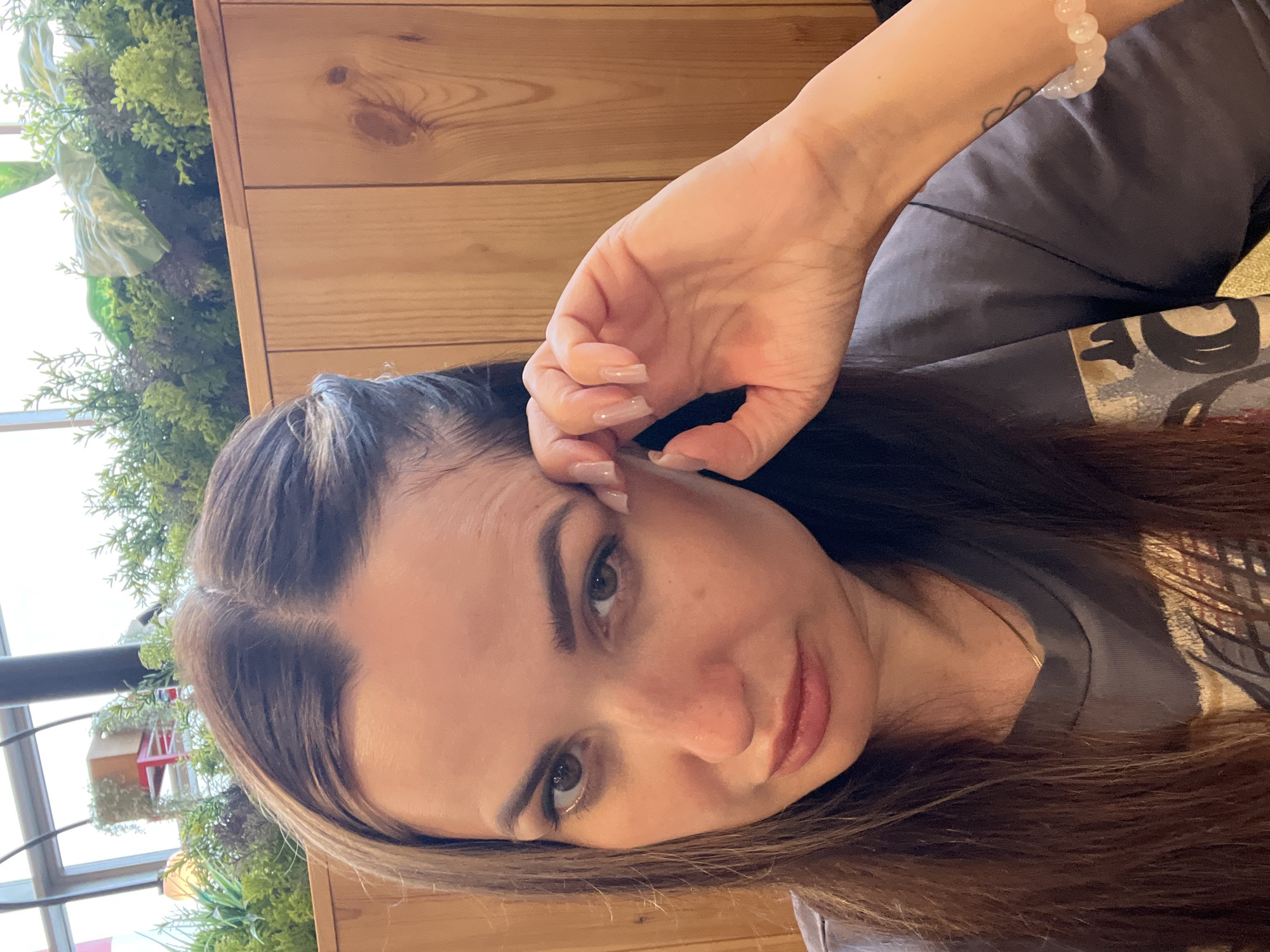 A medium shot captures a woman from the shoulders up, with her head tilted slightly. She is facing the camera, with her right hand resting on her forehead, obscuring part of her eye. Her brown hair is long and falls around her shoulders, with a small section pulled back on the left side of her head. She has brown eyes, dark eyebrows, and is wearing a gray t-shirt. The background consists of wooden paneling and a planter filled with green plants, suggesting an indoor or covered outdoor setting. The lighting appears to be natural, coming from above and to the left.