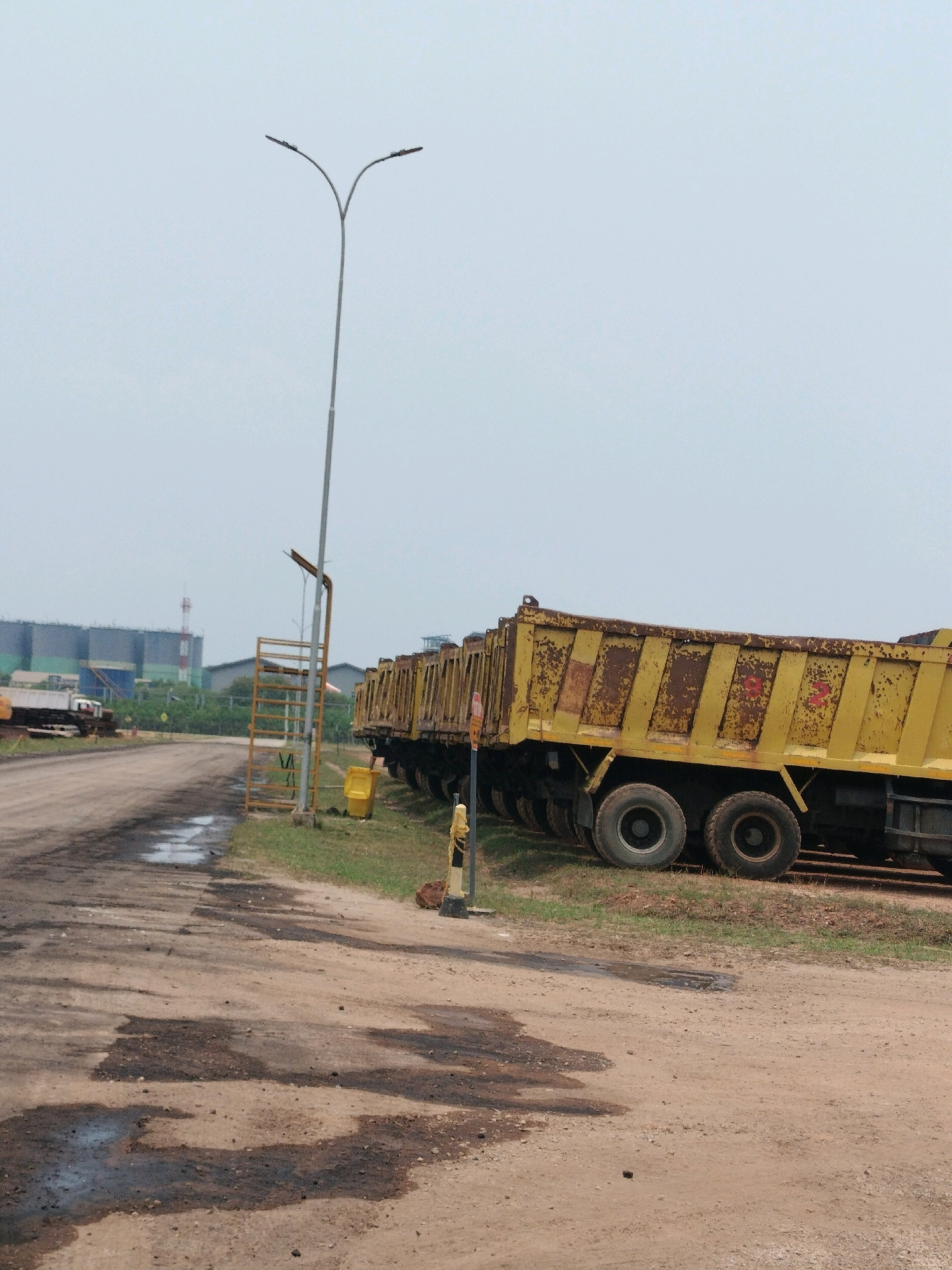 The image captures a scene in what appears to be an industrial or construction area, possibly located near the Batamindo Industrial Park in Batam, Indonesia, given the context of industrial infrastructure. The foreground is dominated by a dirt road, showing patches of wetness and dark stains, suggesting recent rain or leaks. To the right, a line of parked yellow dump trucks, some appearing weathered and rusted, are lined up. These trucks have large tires and their beds are visible, some with visible numbers like "82" painted on them.
In the mid-ground, a tall, modern streetlamp with two lights stands on the left side of the road, its pole reaching high into the overcast sky. Adjacent to the lamp post is a yellow metal ladder-like structure. A yellow trash bin is visible near the base of the lamp post. Further in the background, beyond a strip of green grass and trees, are large industrial buildings with multiple stories, some appearing to be storage tanks or silos, with a prominent red and white striped tower visible behind them. The overall atmosphere is industrial and somewhat desolate, with a clear sky indicating daylight. There are no people visible in the image.