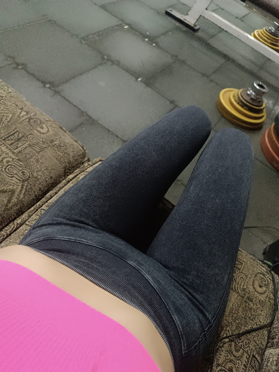 The image shows a first-person perspective looking down at a person's lap and surroundings. The subject is wearing dark blue or grey form-fitting pants or jeans, with a bright pink top visible around the midriff. The skin between the top and pants is visible.
The person appears to be sitting or resting on a patterned brown and tan fabric surface, possibly a couch or bench, which occupies the left and bottom right portions of the frame. The fabric has an intricate, somewhat abstract design.
In the background, there is a dark grey floor, likely rubber gym flooring, composed of square tiles. On this floor, various gym equipment is visible, including several yellow and reddish-brown weight plates stacked on a stand or bar. A white metal structure, possibly part of a weight rack or exercise machine, is also partially visible in the upper right.
The scene suggests an indoor gym or fitness environment, with the person taking a break or observing the surroundings. No specific time of day, weather, or emotions are discernible, and no text is visible.
