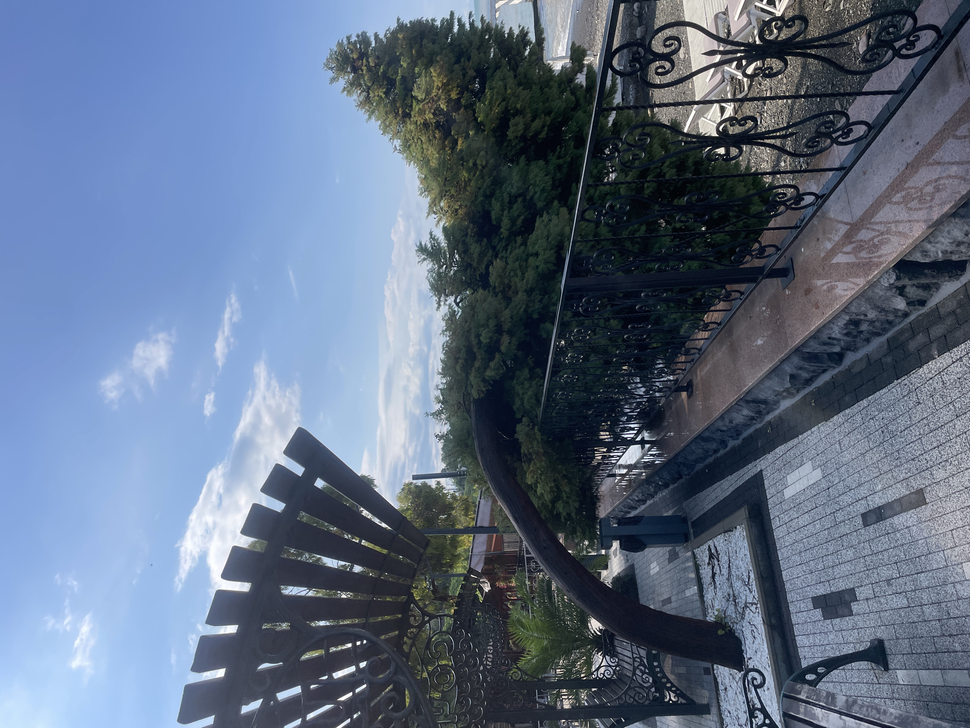 The image depicts an outdoor area with a view of the sea and trees, likely in Sochi, Russia. In the foreground, a wooden pergola-like structure is visible, along with a decorative metal fence. Beyond the fence, dense green foliage from trees obscures much of the background. A pathway made of interlocking pavers leads through the area. The sky is blue with scattered white clouds. The sea is visible in the distance, with a shoreline made of dark pebbles. There are no people visible in the image. The scene appears to be a tranquil outdoor seating or promenade area.