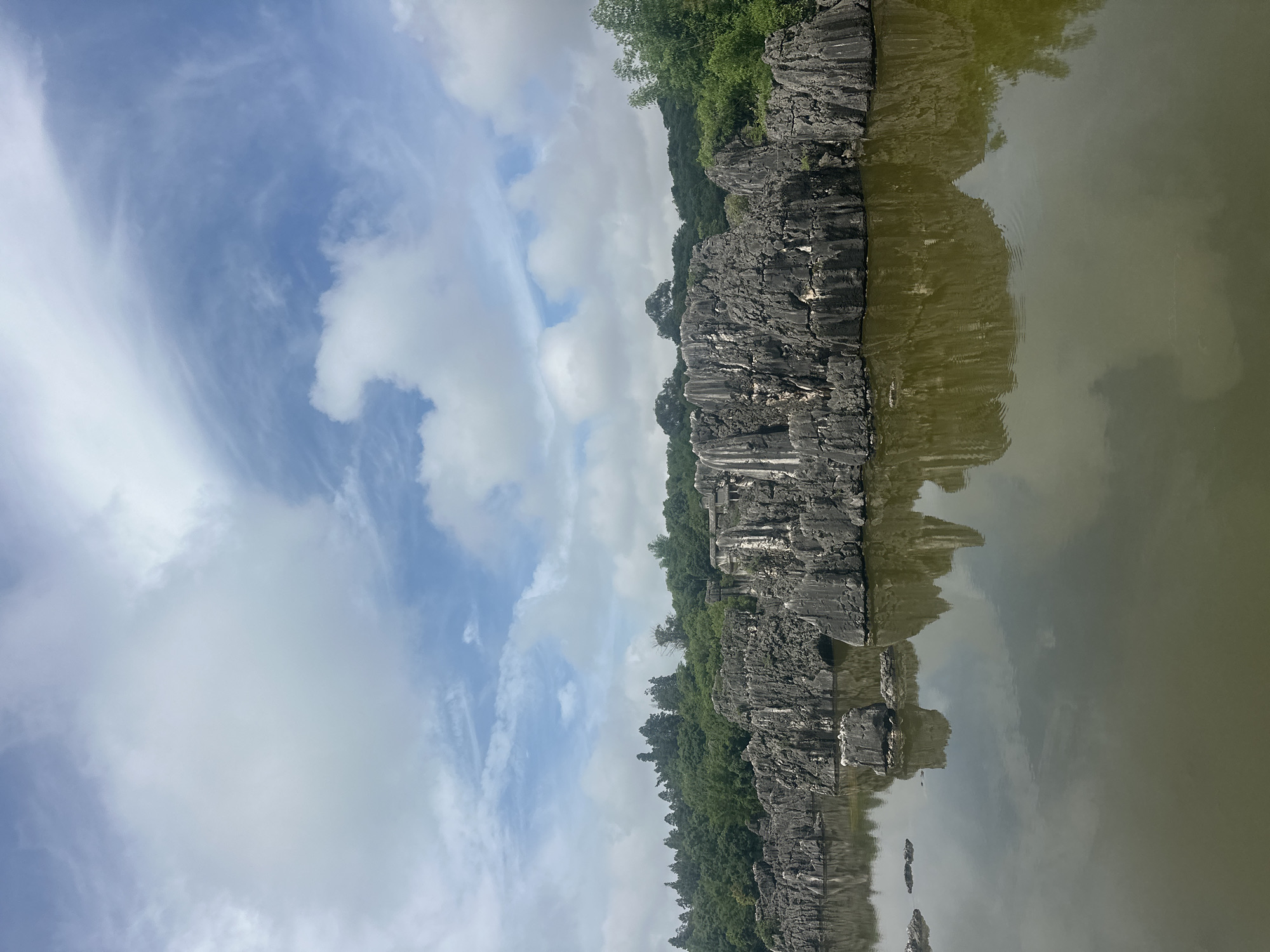 The image depicts the Stone Forest (Shilin) in Kunming, China. A body of calm, dark green water occupies the foreground, reflecting the jagged, grey limestone rock formations that rise from its surface. These formations, characteristic of karst topography, are sharp and columnar, creating an otherworldly landscape. Beyond the rocks, a dense band of lush green trees forms the middle ground, creating a stark contrast with the stone formations. The background is filled with a dynamic sky of white and grey clouds against a bright blue expanse. No people or animals are visible in the scene.