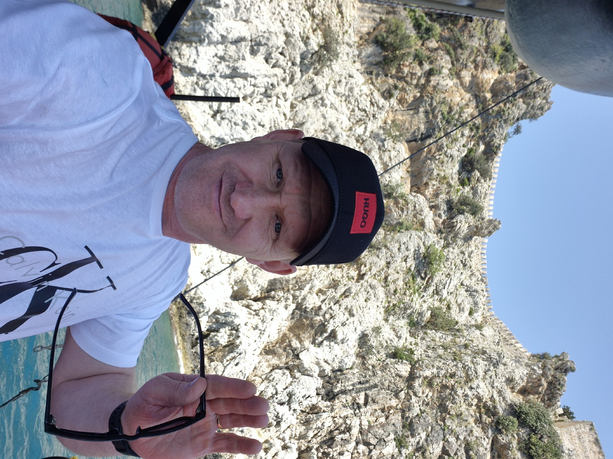 A medium shot depicts a middle-aged male on what appears to be a boat on the water. He is wearing a white t-shirt and a black baseball cap with a red "HUGO" label. He holds eyeglasses in his hand.
Behind the man, a rocky cliff face extends upwards, partially obscuring a blue sky. The cliff shows signs of vegetation.
Based on the cliff formation and the water, this scene appears to be from the city of Alanya, Turkey, near the Alanya Castle.