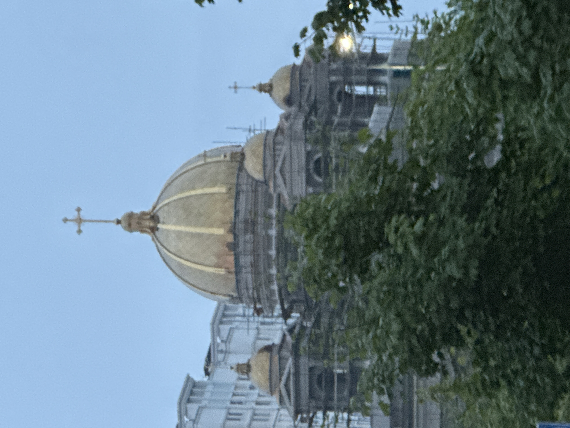 The image shows the gold domes of a cathedral with crosses on top. The cathedral is in Podolsk, Russia. The sky is overcast and the trees in the foreground are green. The cathedral appears to be a grand and ornate structure, possibly a historic landmark or a prominent religious building in the city. There are no people visible in the image. The overall scene suggests a calm and somewhat somber atmosphere due to the cloudy weather.