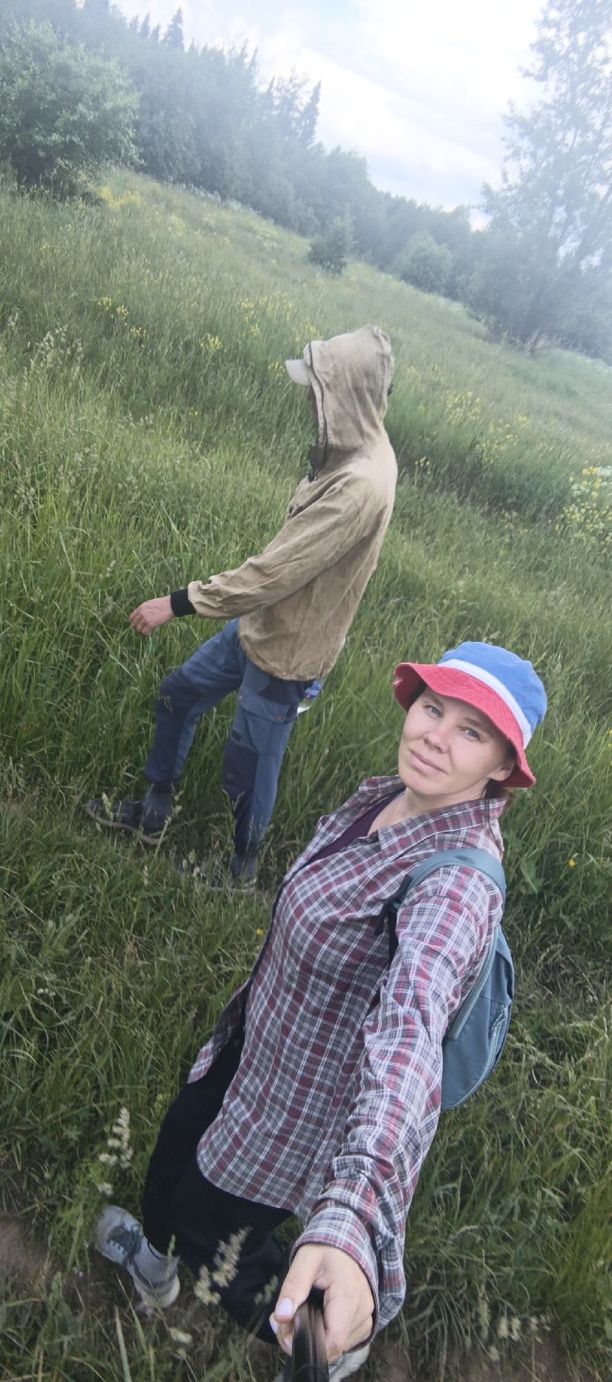 In a grassy, outdoor environment, likely near Garevaya, Russia, two individuals are present. In the foreground, a woman wearing a red and white checkered shirt, a blue and red bucket hat, and a backpack is holding a selfie stick, capturing the image. Her facial features are visible, and she appears to be looking at the camera. Behind her, further up a grassy incline, a person wearing a hooded jacket in a neutral color and blue pants is visible from the side. This individual is facing away from the camera, with their hood up, and their body is angled towards the left of the frame. The background consists of dense green grass with scattered yellow wildflowers and a treeline further up the hill. The lighting suggests it is daytime, possibly overcast.
