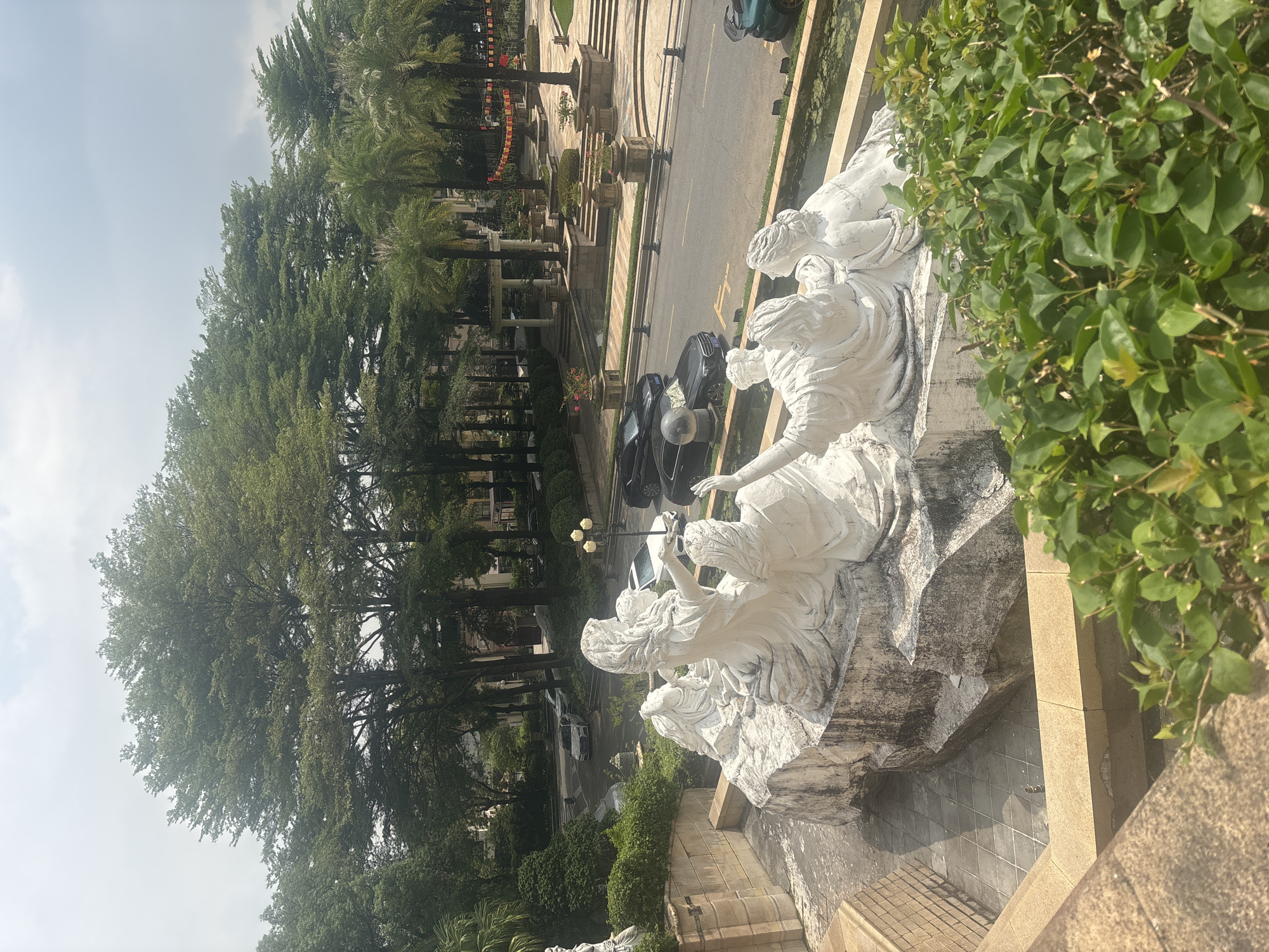 In the Nanhai District of China, a scenic outdoor setting is captured, featuring a white sculpture of several figures in dynamic poses, appearing to be in motion. The sculpture rests on a rough, rocky base, with the figures draped in flowing garments. In the background, a paved road is visible with several cars, including a white sedan and black sedans, parked and driving. The environment is lush with various trees, including tall, slender palm trees and broader, leafy trees. The ground is a mix of paved surfaces and manicured green spaces, with some ornamental plants in planters. The sky is overcast, suggesting a cloudy day. The perspective is from a slightly elevated position, looking down at the sculpture and the scene beyond.