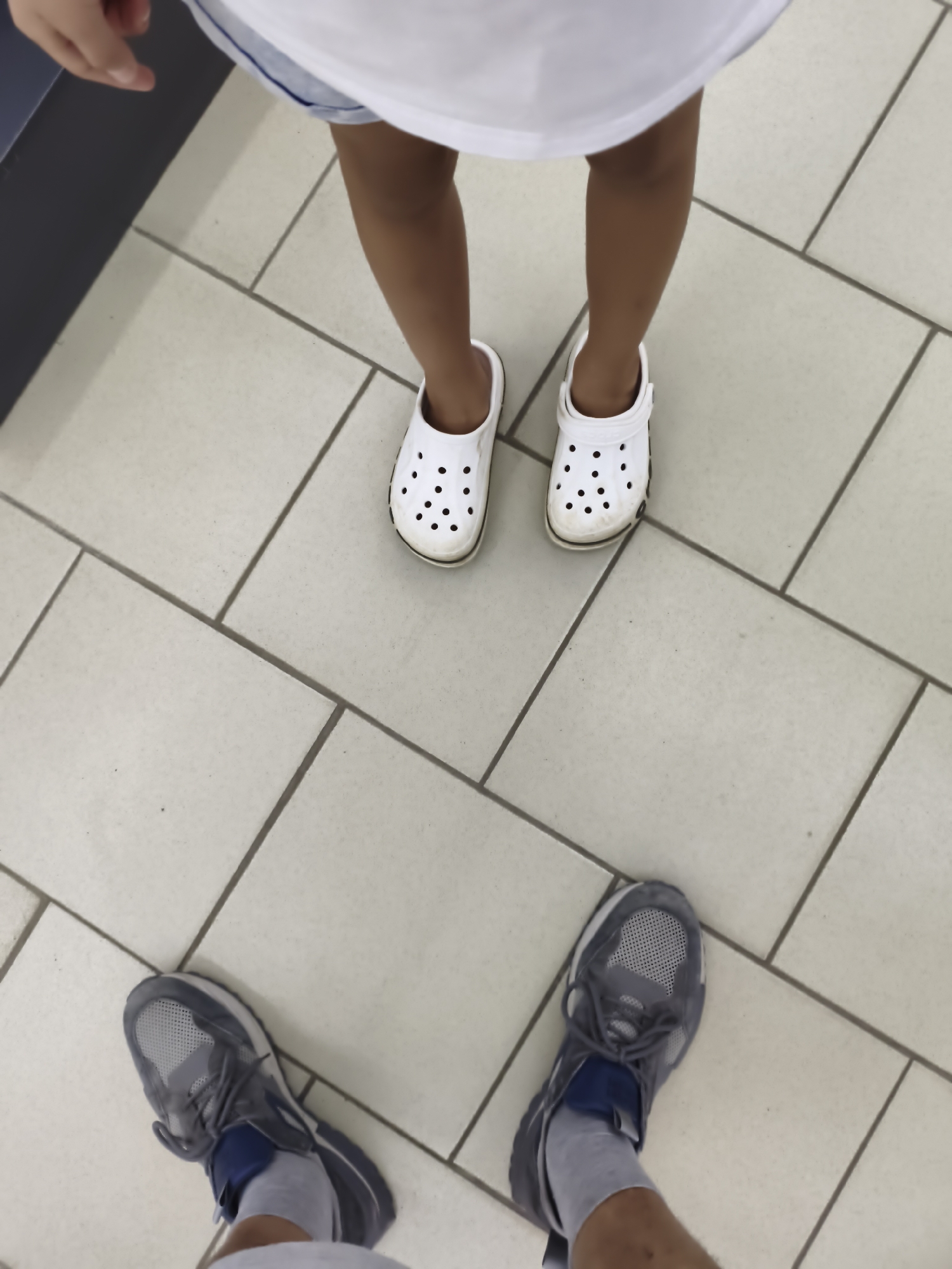 The image is a ground-level, overhead view of two pairs of feet standing on a tiled floor. The feet on the upper portion of the image belong to a child, wearing white clogs with black ventilation holes. The child's legs are visible, wearing what appears to be a white shirt and light blue shorts. The feet on the lower portion of the image belong to an adult, wearing grey athletic shoes with blue accents and grey socks. The adult's legs are also visible. The environment appears to be indoors, with a floor composed of large, square, light-colored tiles with dark grout lines. The lighting suggests an indoor space, possibly a commercial establishment or public building. The context of Aktobe, Kazakhstan, does not provide specific visual cues within the image itself, but implies the geographical setting of the photograph.