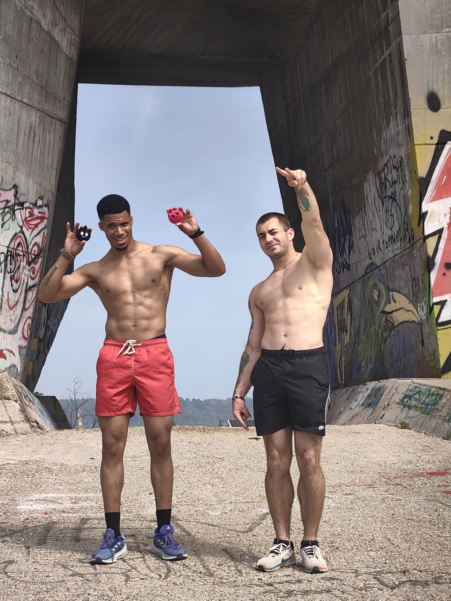 The image displays two shirtless men standing on a light-colored ground composed of gravel and small pebbles, beneath a large concrete structure. This structure, characterized by a wide arch and extensive, colorful graffiti, is consistent with the areas under or near the Asparuhov Bridge in Varna, Bulgaria. Beyond the arch and structure, a hazy light-blue sky meets distant hills sparsely covered with dry vegetation.
The man on the left, with a dark complexion, possesses a muscular physique and short dark curly hair. He is smiling directly at the camera. He wears red swim shorts with white drawstrings, black socks, and blue athletic shoes with white soles. In his raised left hand, he holds a small black plush toy with a simple face, and in his raised right hand, he holds a matching red plush toy. Tattoos are visible on his left bicep and forearm, and he wears a bracelet on his left wrist.
The man on the right, with a light complexion, also exhibits a muscular build and has short dark hair with light facial stubble. He wears black athletic shorts featuring a white stripe and a Nike logo on the left leg, paired with light-colored athletic shoes that have black laces and subtle pink/orange accents on the soles. His right arm is raised, with his index finger pointing upwards and to the right. A colorful tattoo, appearing to be a plant or leaf design, is visible on his right bicep. Tattoos are also present on his left arm, and he wears a bracelet on his left wrist. His left hand is partially obscured by his shorts.
The concrete walls of the overhead structure are extensively covered in various spray-painted tags, lettering, and graphic art, including a prominent green bird-like creature with a yellow beak on the right support column. The ground underfoot appears rough and unpaved.