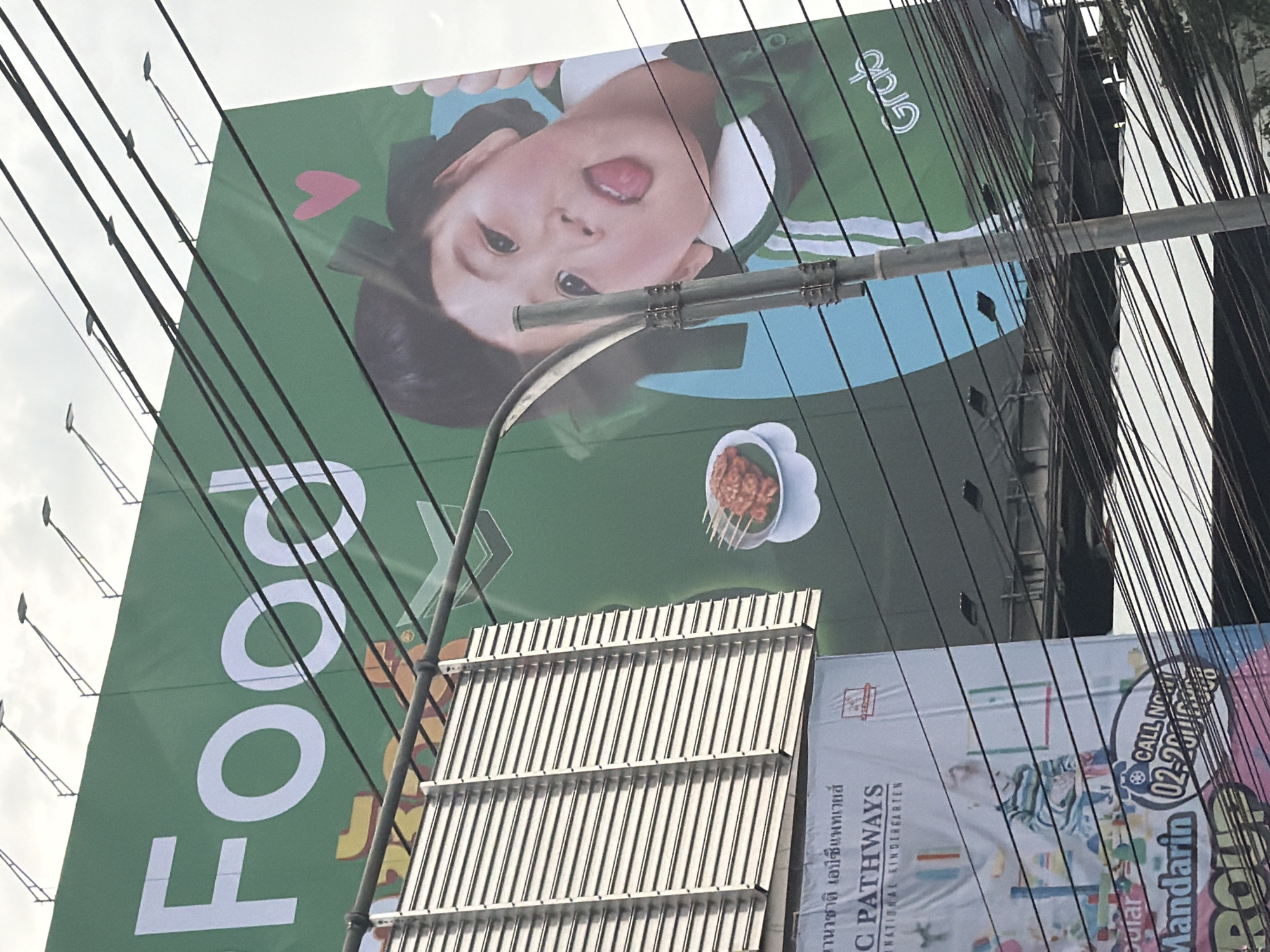 This image, captured in Bangkok, Thailand, features a large green billboard advertising "Food". The billboard displays a close-up of a young child with their tongue sticking out, a pink heart, and a plate of grilled skewers. The Grab logo is visible on the right side of the billboard. Below the main billboard, another advertisement for "C PATHWAYS INTERNATIONAL KINDERGARTEN" is partially visible, showing a child playing with building blocks and the text "Mandarin" and a phone number. The scene is obscured by a dense network of power lines. The sky is overcast.