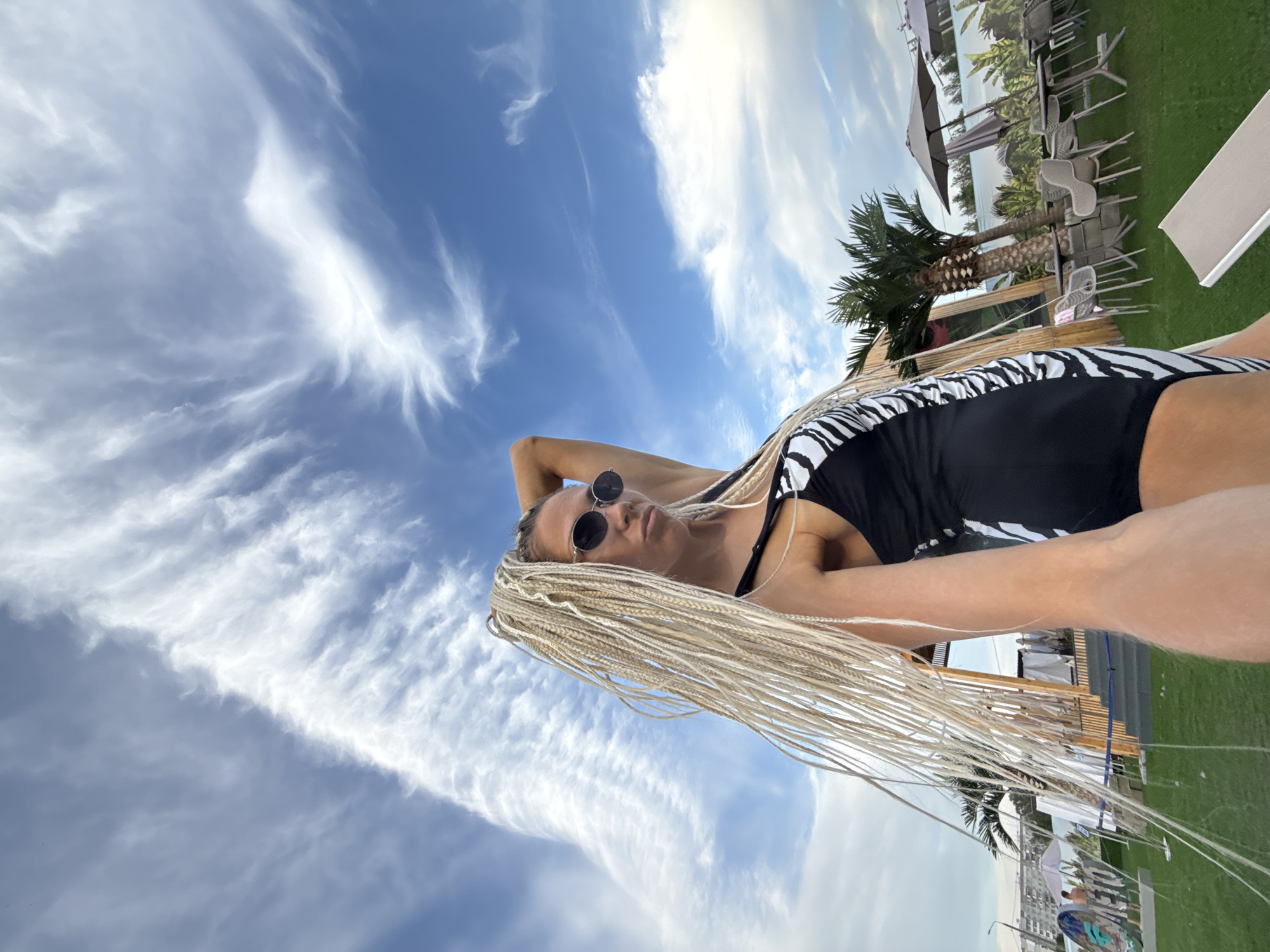 The image shows a woman lying down outdoors, with her head tilted back, looking up at the sky. She has long, blonde braided hair and is wearing a black swimsuit with a zebra print. She also has on sunglasses. The sky is a bright blue with dramatic, wispy white clouds.
In the background, there is a grassy area with several outdoor tables and chairs, some of which are under umbrellas. Palm trees are visible, suggesting a warm climate, however, the location context given is Yaroslavl, Russia. There appears to be a body of water and some buildings in the distance. The image appears to be taken in the daytime, with bright sunlight. The overall mood seems relaxed and summery. No visible text is present in the image.