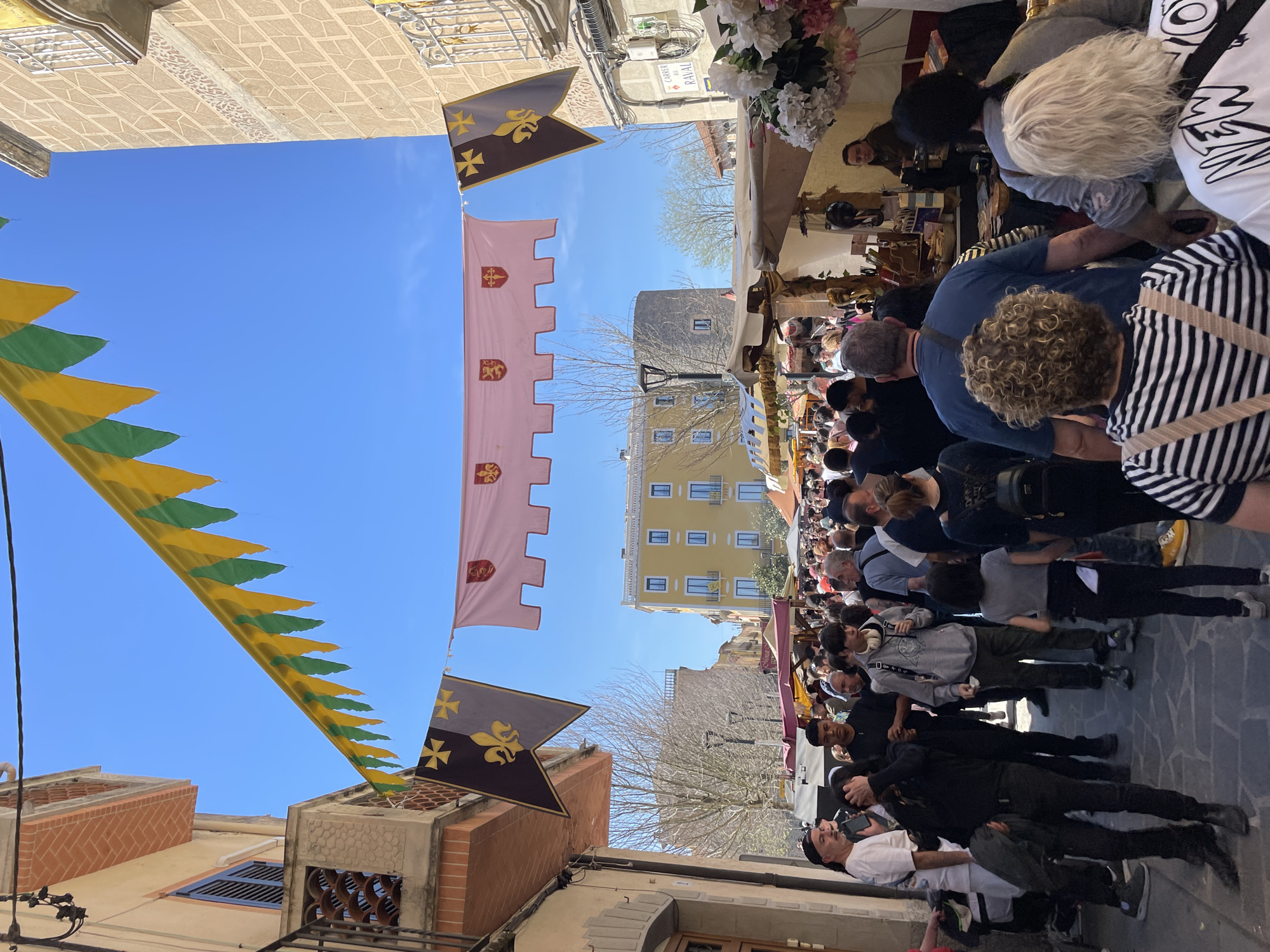 The image shows an outdoor street market scene in Hostalric, Spain. A crowd of people walks down a narrow street lined with market stalls and decorated with medieval-style banners and pennants. Banners hang on either side of the street, including one with a fleur-de-lis and a cross symbol on a dark background, and another with a castle tower design on a light pink background. Yellow and green pennants are also visible. The street is lined with buildings of different heights and colors. The sky is clear and blue. The market stalls appear to have various wares for sale. Some people are looking at the stalls, while others are simply walking through the crowd.