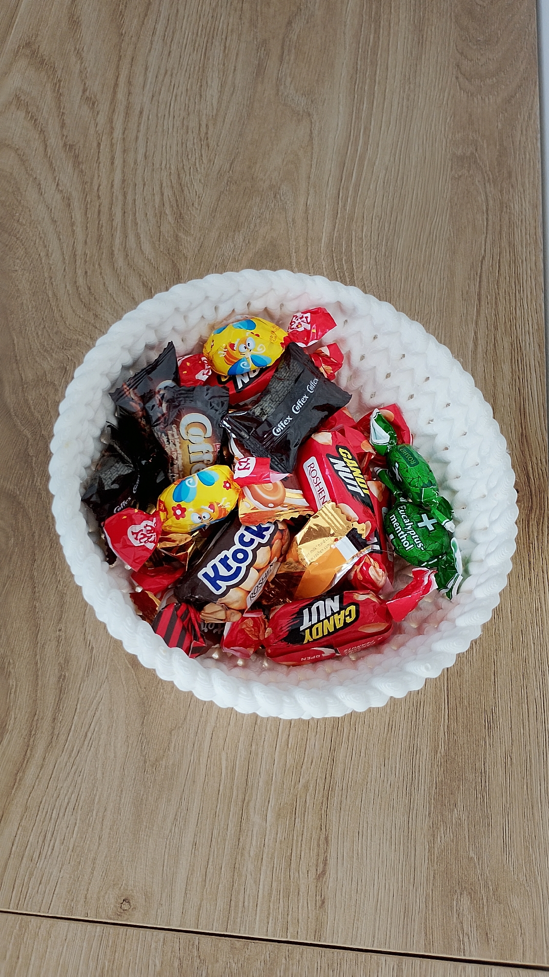 A white, crocheted bowl filled with assorted wrapped candies sits on a light brown wooden surface. The candies are in various wrappers, including red, black, gold, and green. Some visible brands and wrappers include "Kroft," "Roshen," "Candy Nut," and "Coffex." There is no indication of the location being Smolyan, Bulgaria.
