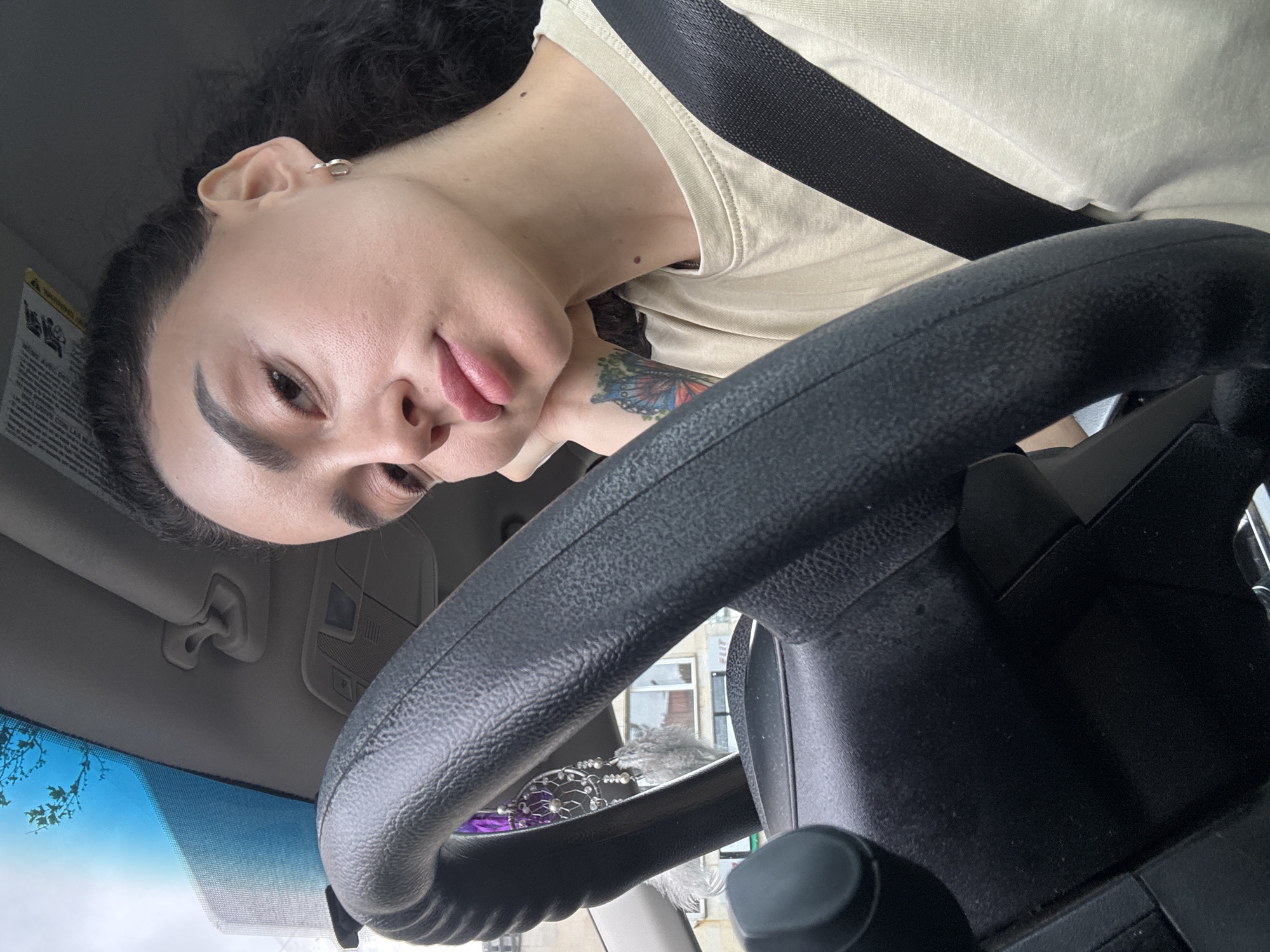A person is seated in the driver's seat of a vehicle, wearing a tan-colored shirt and a black seatbelt. The person has dark hair pulled back, an earring in their left ear, and a butterfly tattoo on their neck. The steering wheel of the car is prominently visible in the foreground. Outside the windshield, through the steering wheel, a building is visible, indicating an urban environment. The sky is overcast. The location cannot be definitively determined as Tbilisi, Georgia, without further visual cues or metadata.