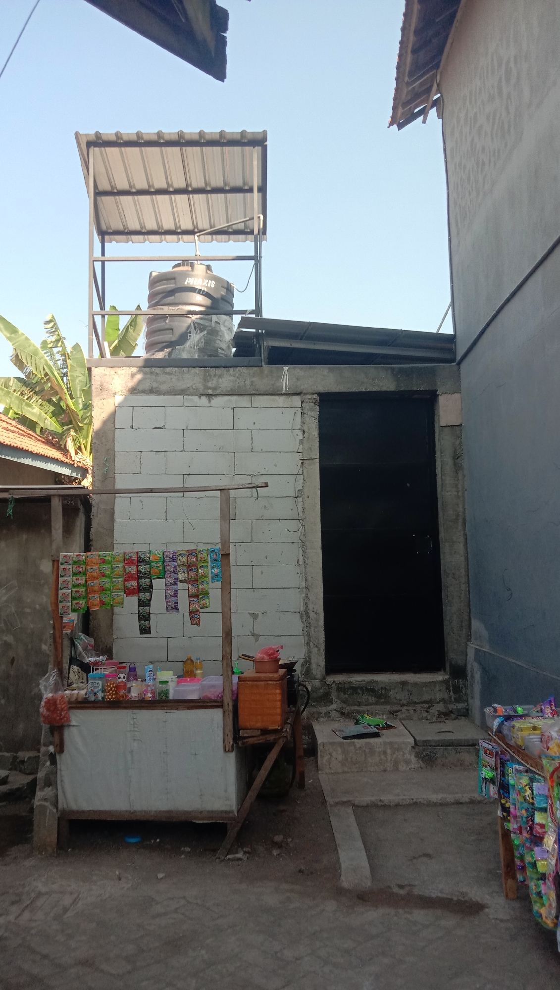 A low-angle outdoor shot depicts a small roadside store and a water tank elevated on a platform in Karang Anyar, Indonesia. The scene is dominated by concrete block construction and corrugated metal roofing. A small stall with a white canvas front displays various bagged snacks hanging from a wooden frame, with additional items and a woven storage bin on its surface. To the right, a tall, black metal door is set into a concrete wall, with concrete steps leading up to it. Above the stall, a large black water tank, labeled "PRAXIS," sits on a metal structure with a corrugated metal roof, partially obscured by a banana plant. The surrounding ground is dirt and gravel. Another display of bagged goods is visible to the far right of the frame.