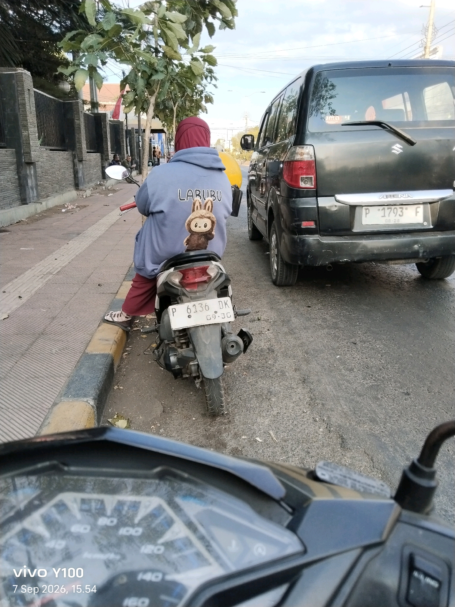 In Situbondo, Indonesia, a person wearing a red headscarf and a grey hoodie with a cartoon rabbit design is seated on a motorcycle. The hoodie displays the text "LABUBU" above the graphic. The person is wearing maroon pants and sandals. The motorcycle's license plate reads "P 6136 DK 09-30". To the right of the motorcycle is a black car, a Suzuki Arena, with the license plate "P 1793 F 06-26". The car appears to be parked or stopped on the asphalt road. The motorcycle is positioned on the edge of a sidewalk constructed with textured grey paving stones and bordered by a curb with yellow and grey markings. Trees with green leaves line the sidewalk, and a grey stone wall is visible in the background. The sky above is partially cloudy. The image is taken from the perspective of another motorcycle, with its dashboard visible in the foreground, displaying the text "vivo Y100" and the date "7 Sep 2026, 15.54".